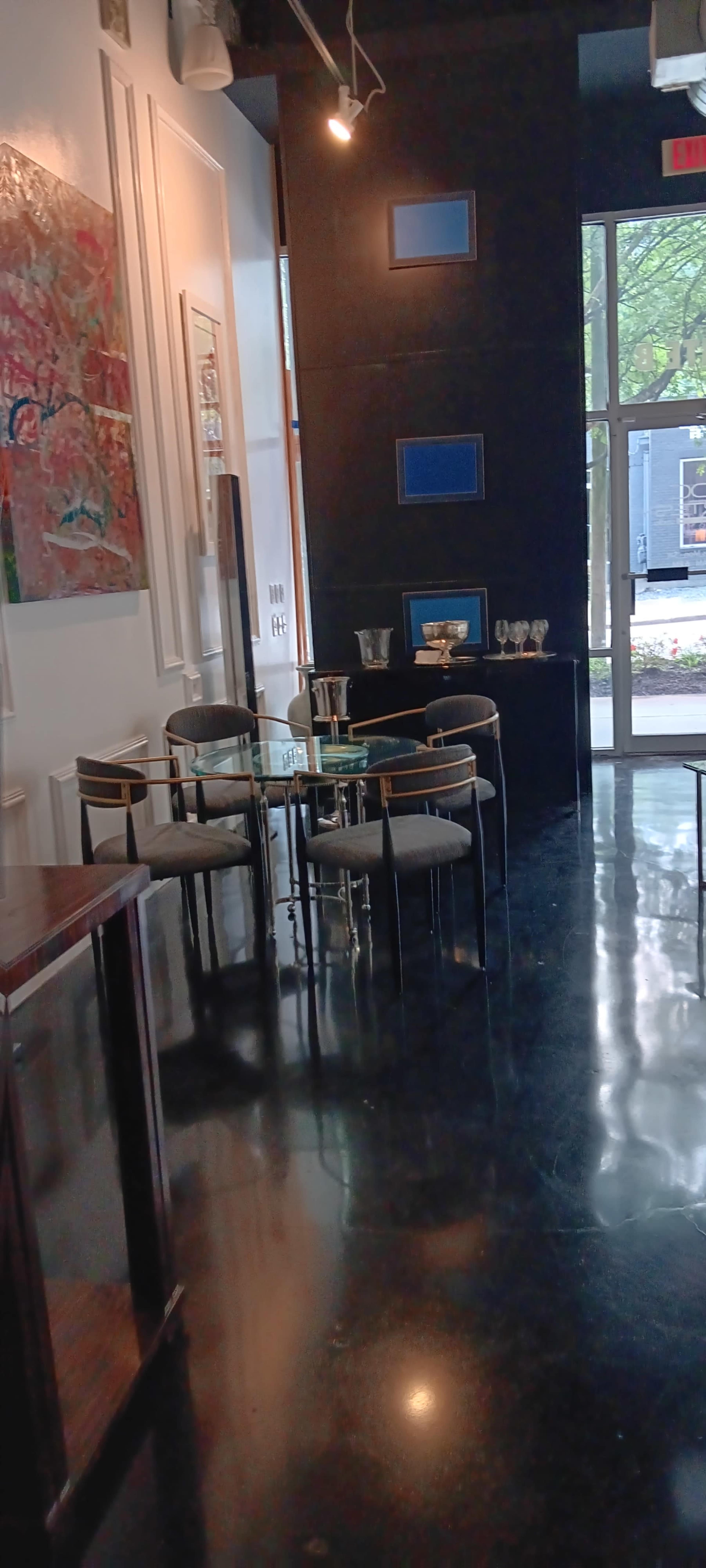 The image shows a modern interior space with a glass dining table surrounded by four upholstered chairs, artwork on the walls, and a large window facing a courtyard.