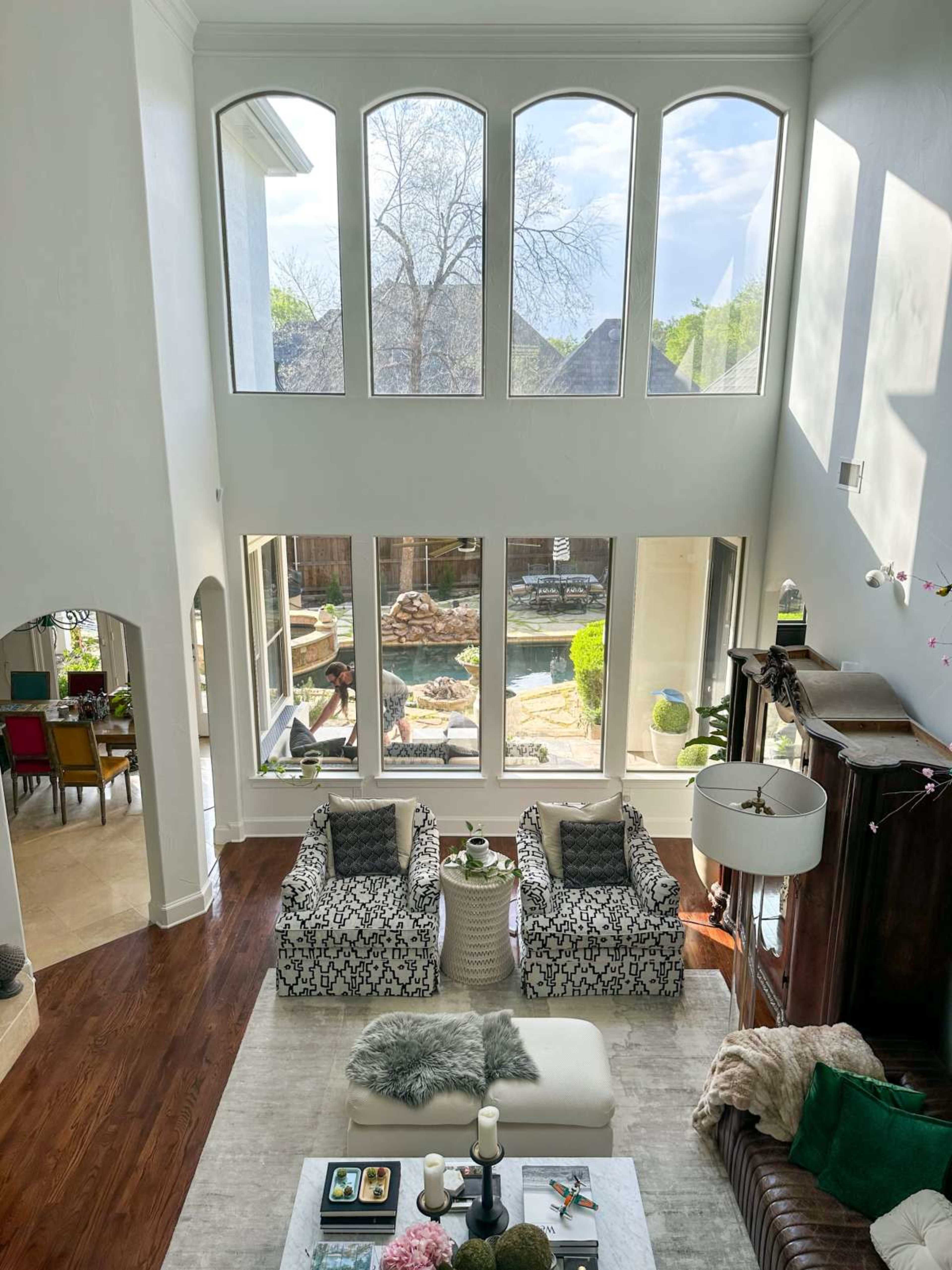 A spacious living room features two patterned chairs facing a coffee table, with large windows offering a view of a backyard pool and garden.