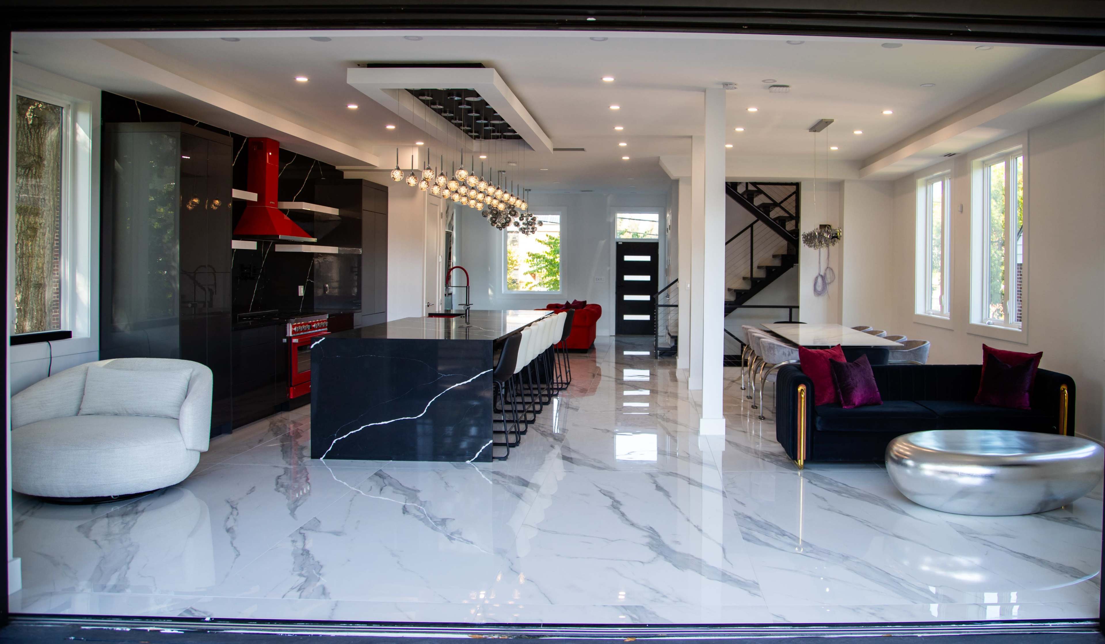 The image shows a modern open-concept living area with a kitchen featuring a large island, sleek appliances, and a dining space, all illuminated by pendant lights and large windows.