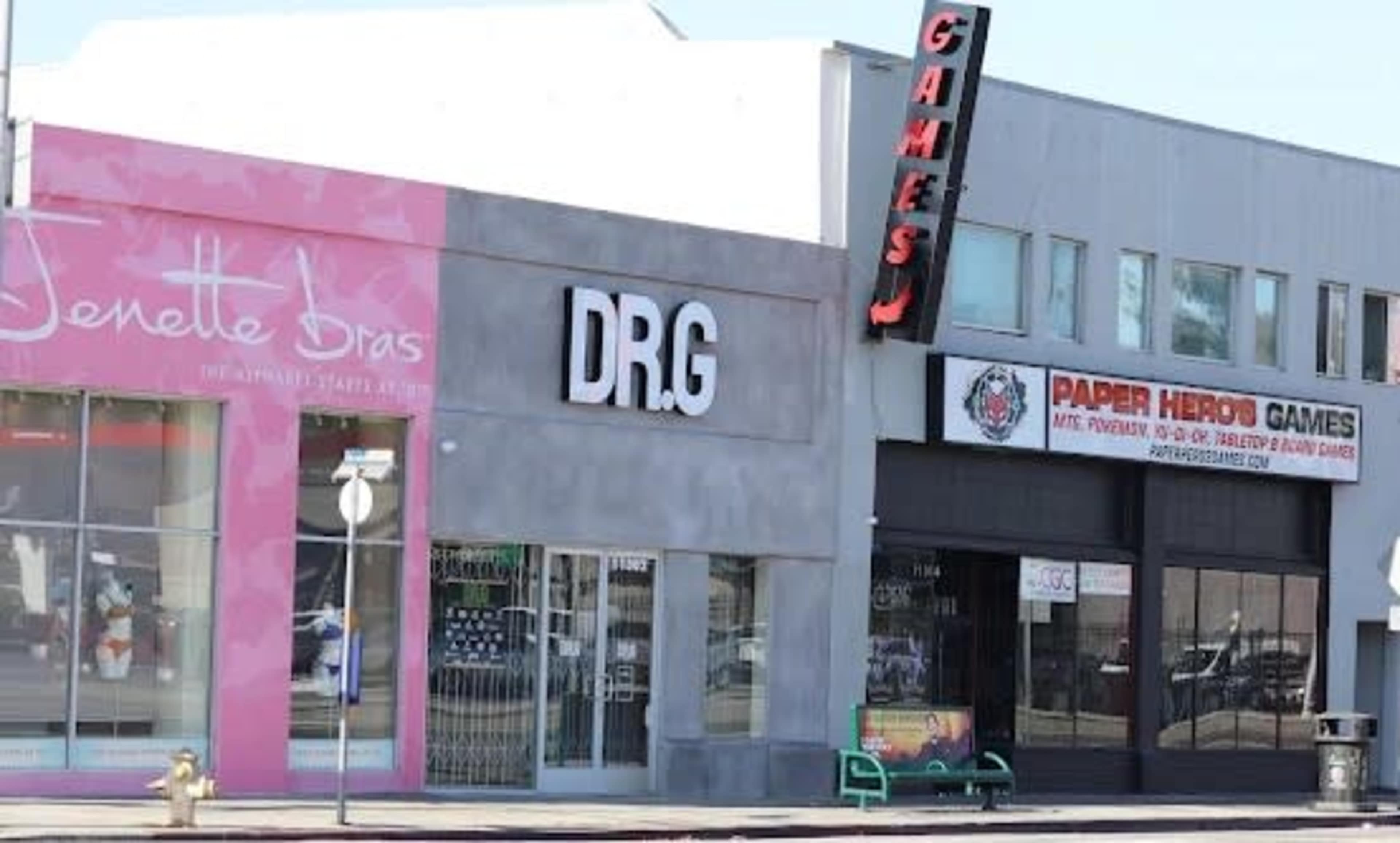 The image shows a street scene featuring two storefronts: one with a pink facade labeled "Jenette Bras" and the other a gray building with "DRG" and "Paper Hero Games" signs.