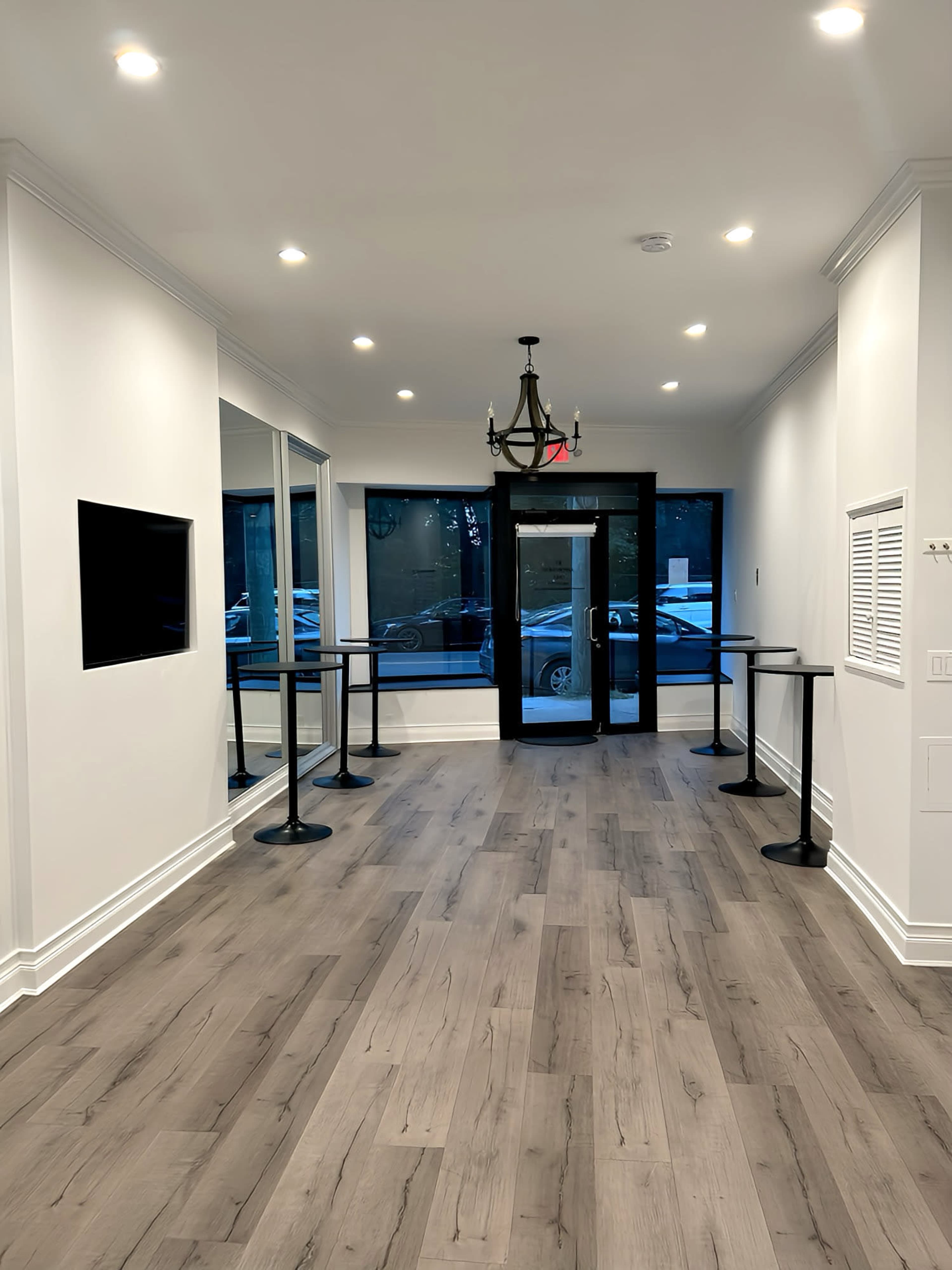 The image shows a modern, well-lit hallway with wooden flooring, featuring a glass door at the end, black standing tables on either side, and a chandelier overhead.