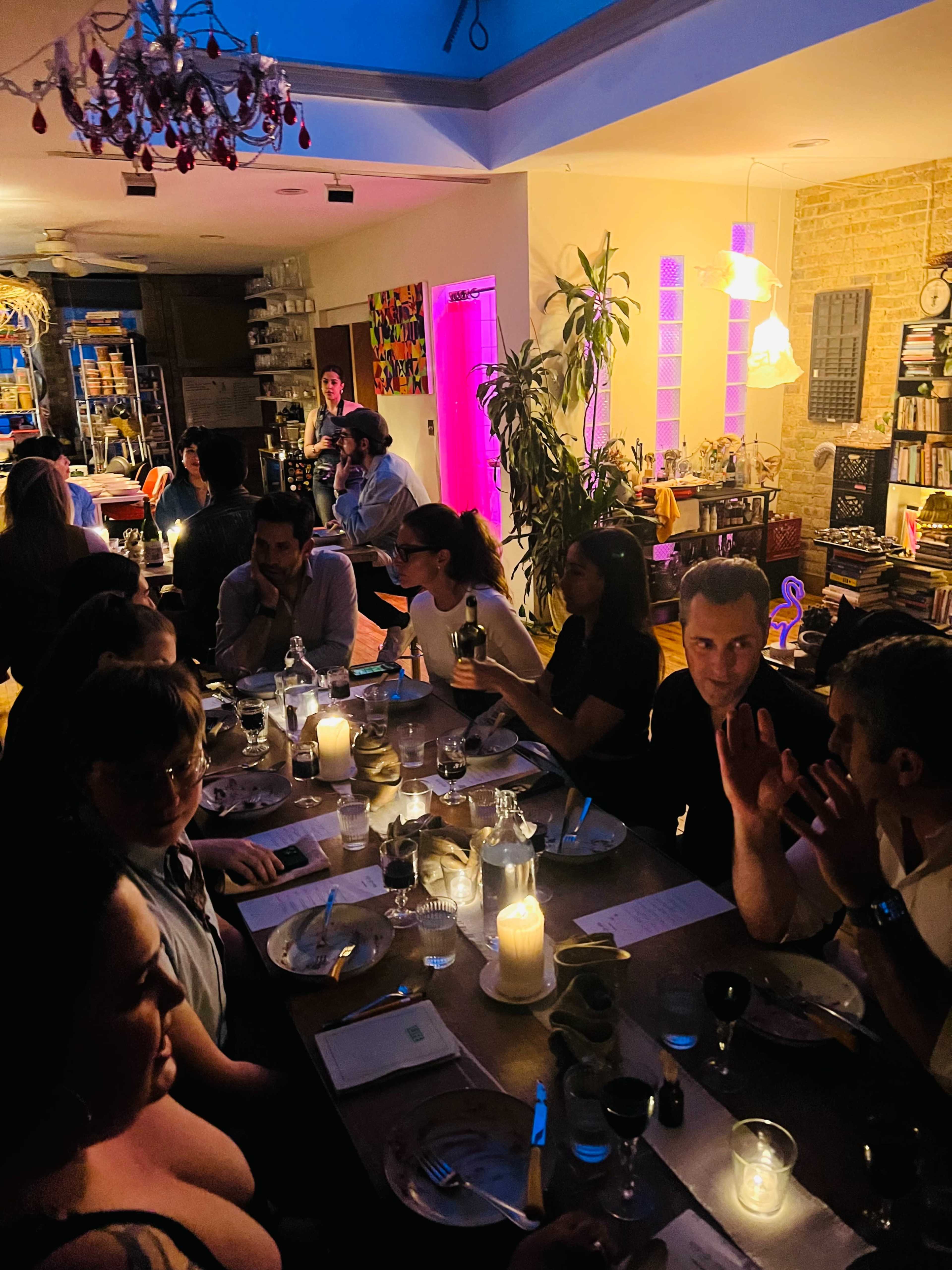 A group of people is seated around a dimly lit dining table, engaged in conversation while candles illuminate the setting.