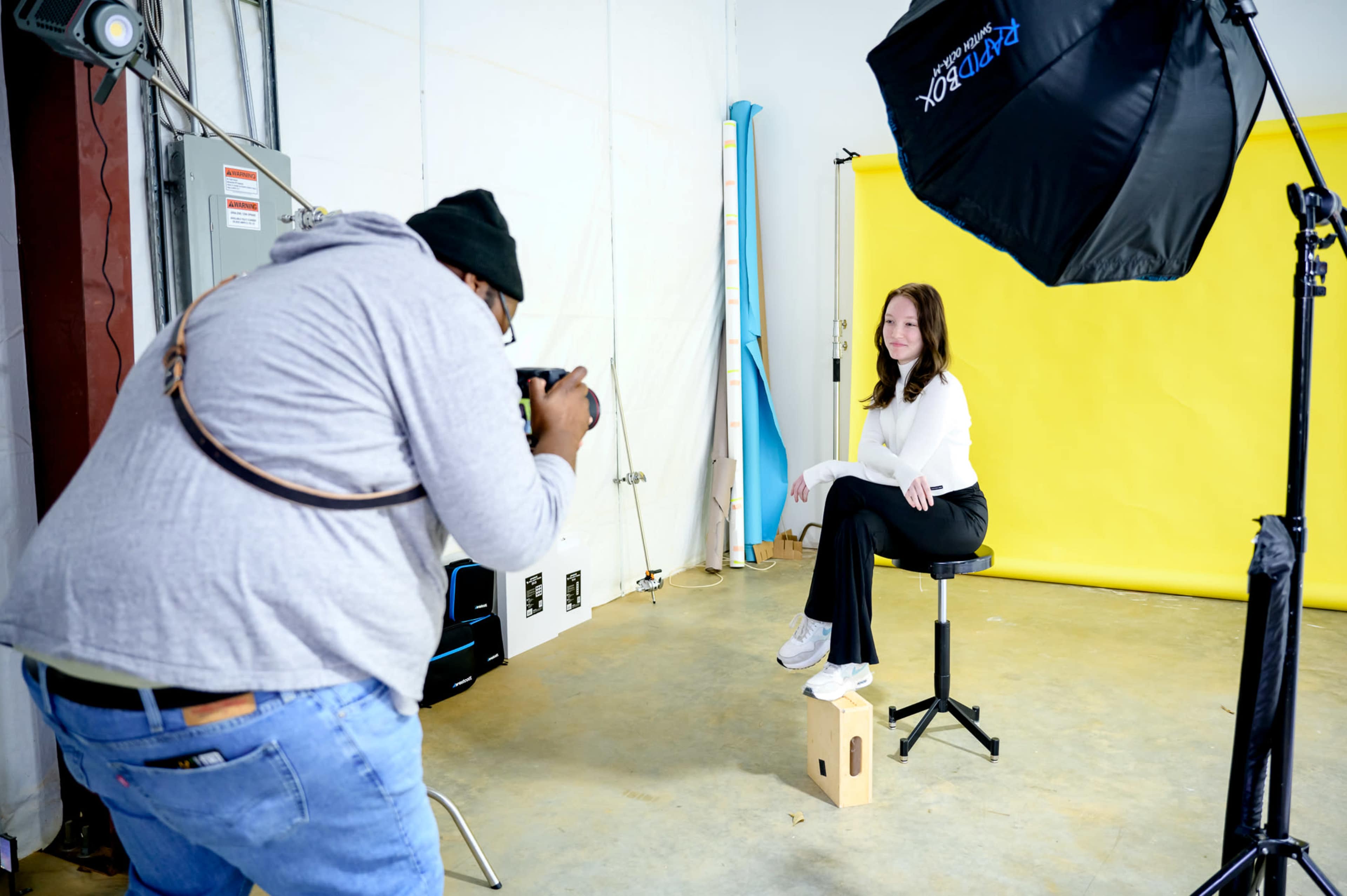A photographer captures a subject posing on a stool in front of a yellow backdrop in a studio.