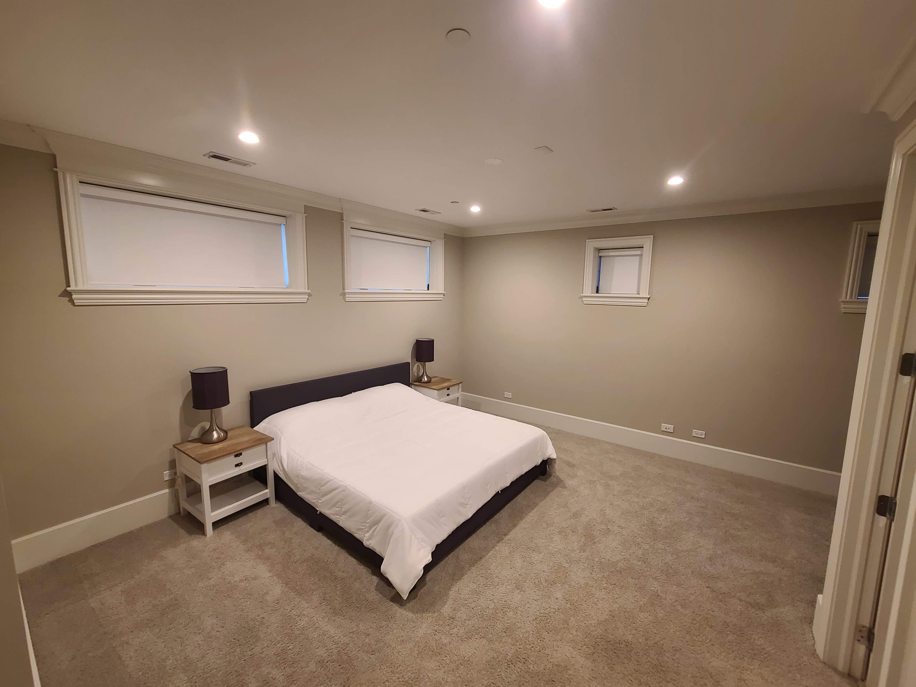 A spacious bedroom featuring a bed with a white comforter, two bedside tables with lamps, and light-colored walls.