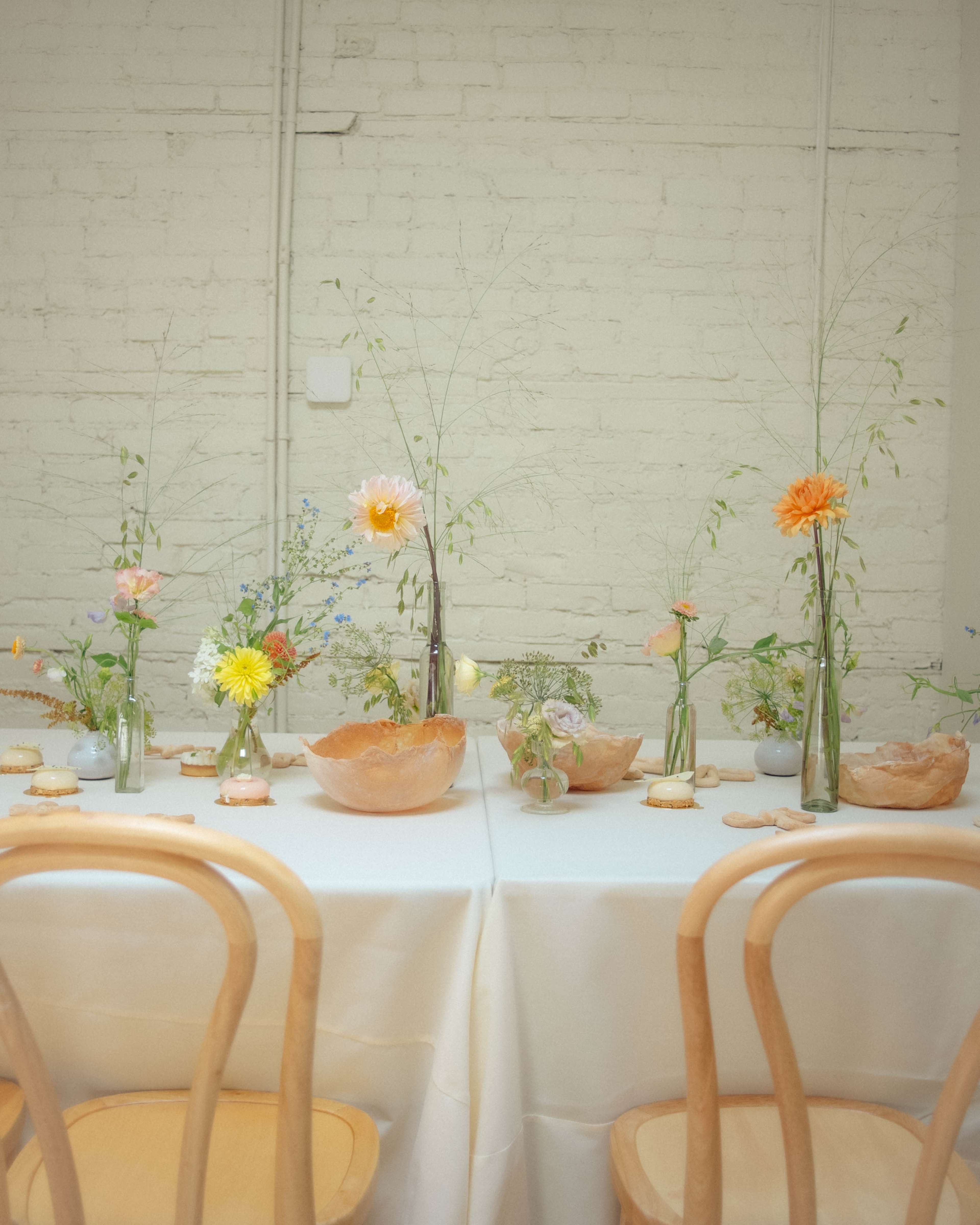 A long table is set with delicate floral arrangements and decorative bowls, surrounded by wooden chairs.
