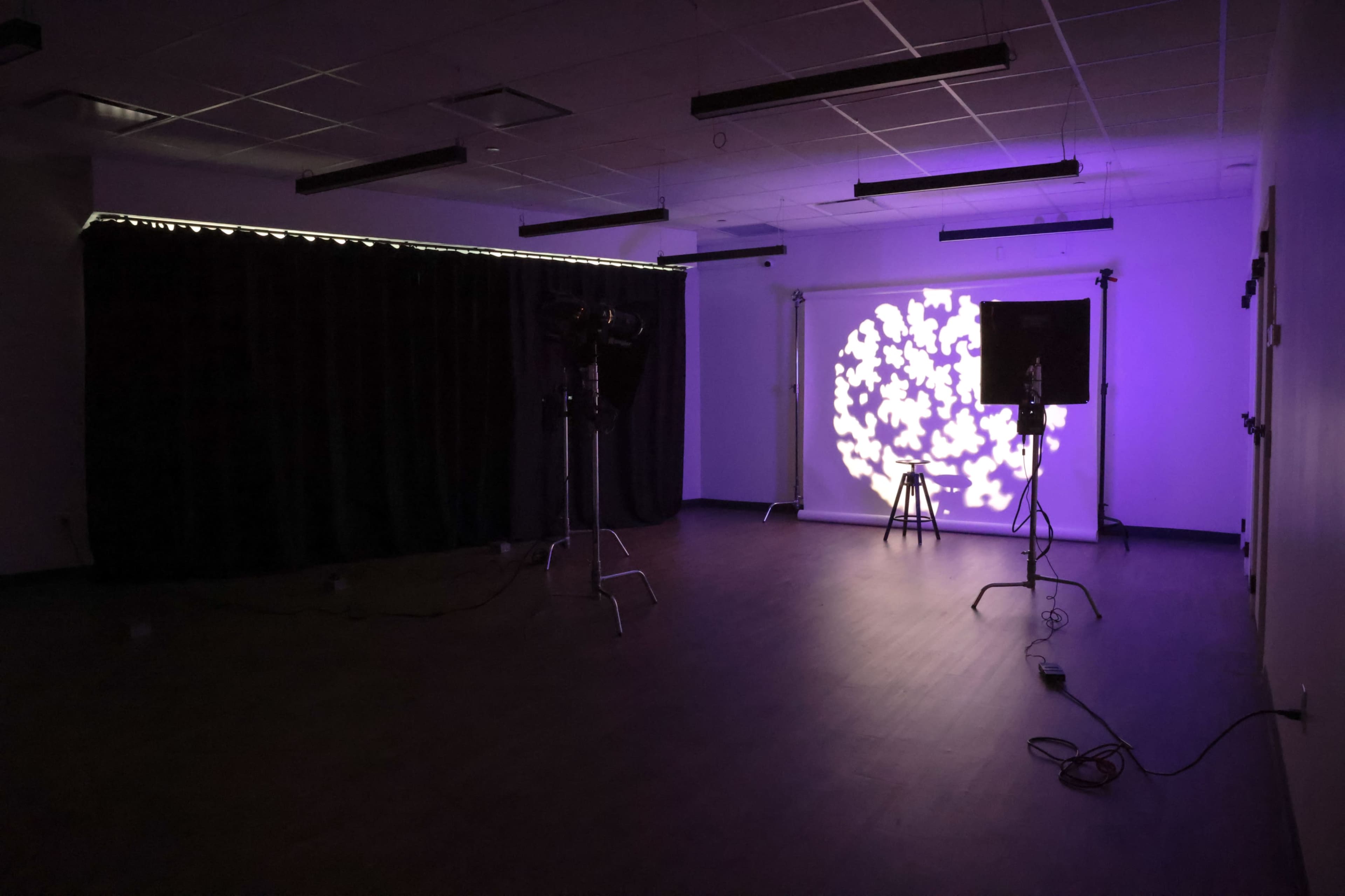 The image shows a dimly lit room with a projection of purple shapes on the wall, accompanied by a light stand and a dark curtain in the background.