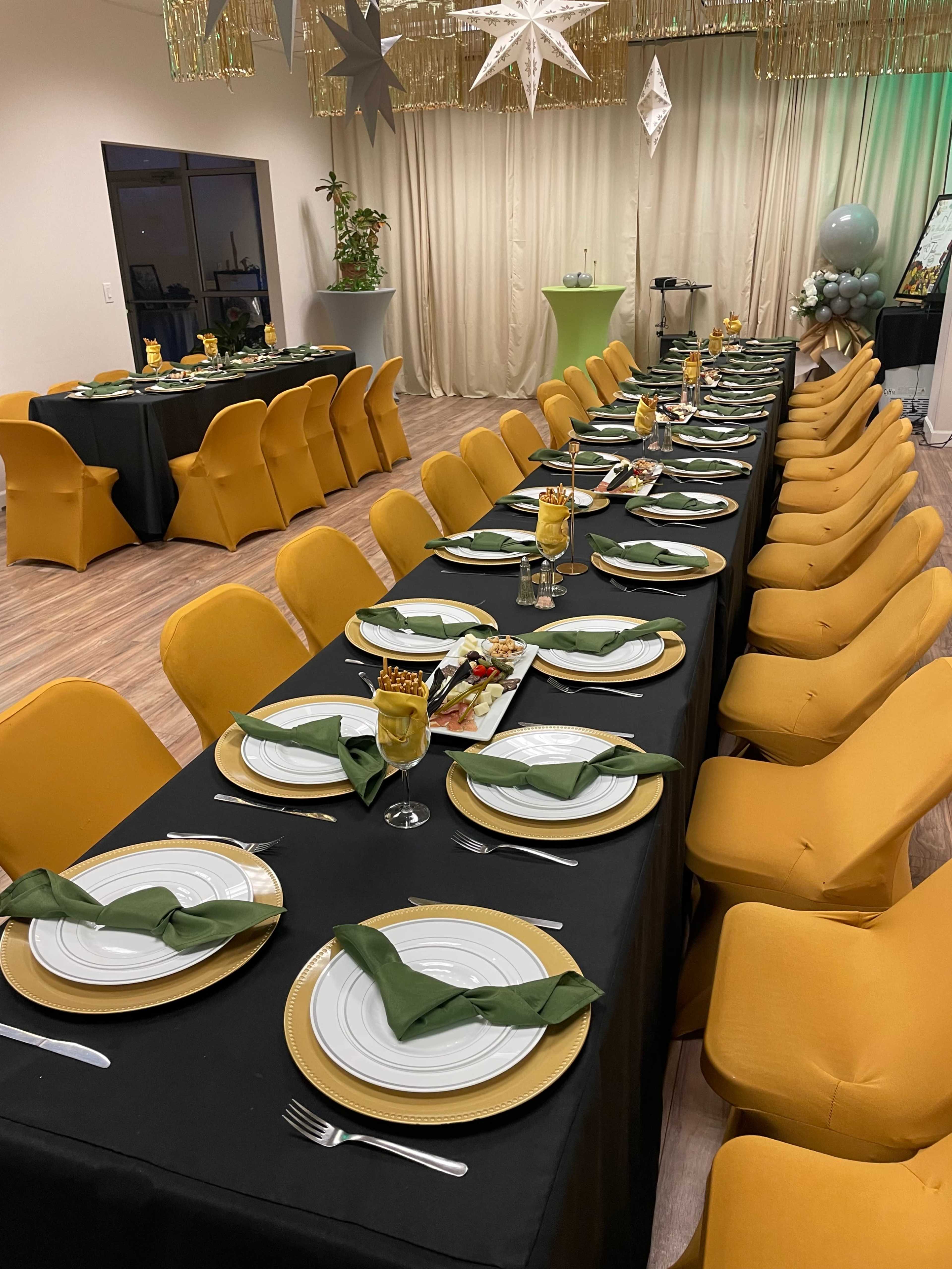 A long dining table is set with green napkins and golden plates, surrounded by yellow chairs in a decorated event space.