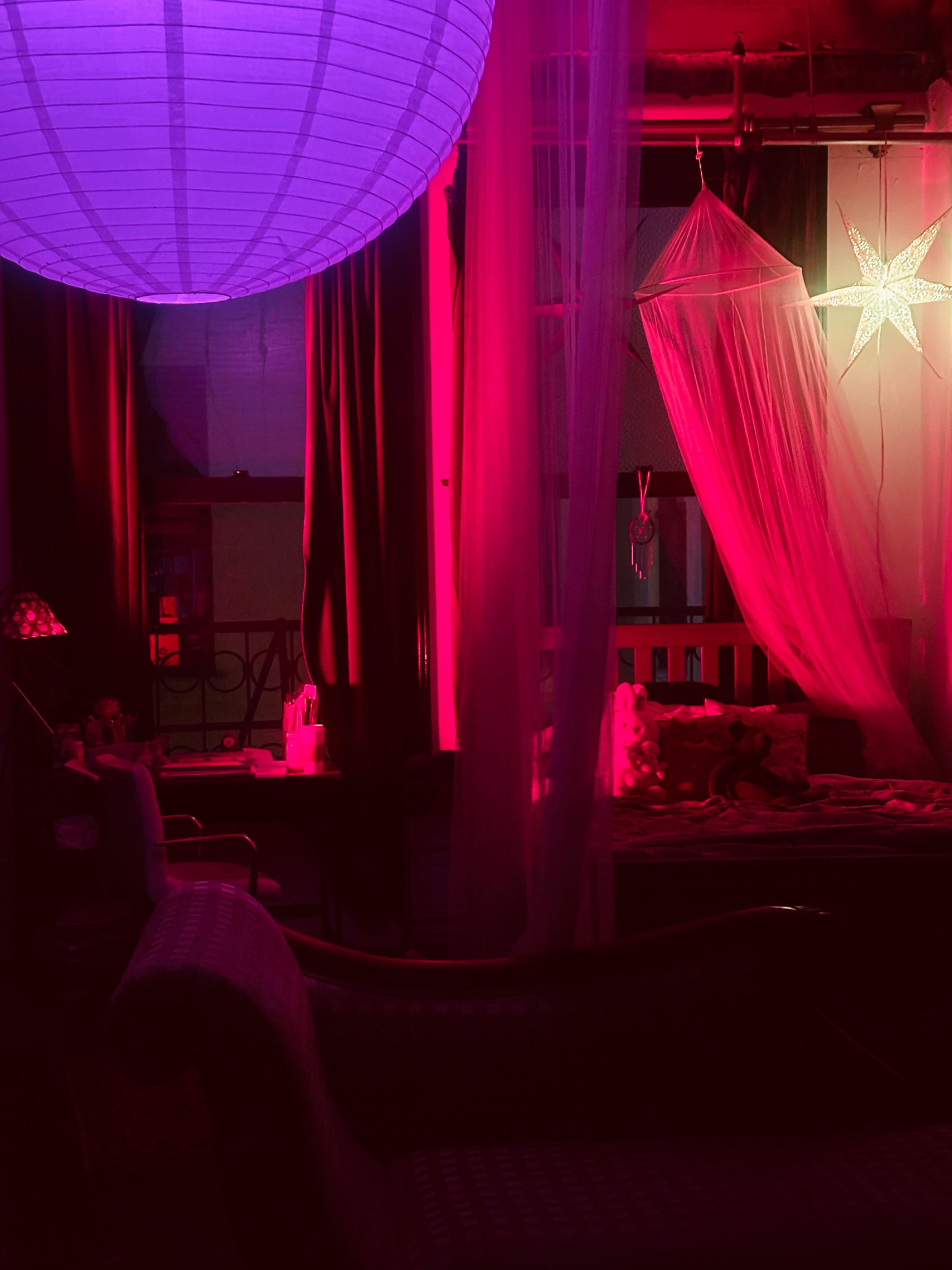 The image shows a dimly lit room with colorful lighting, featuring a bed draped in sheer fabric and a large paper lantern hanging from the ceiling.
