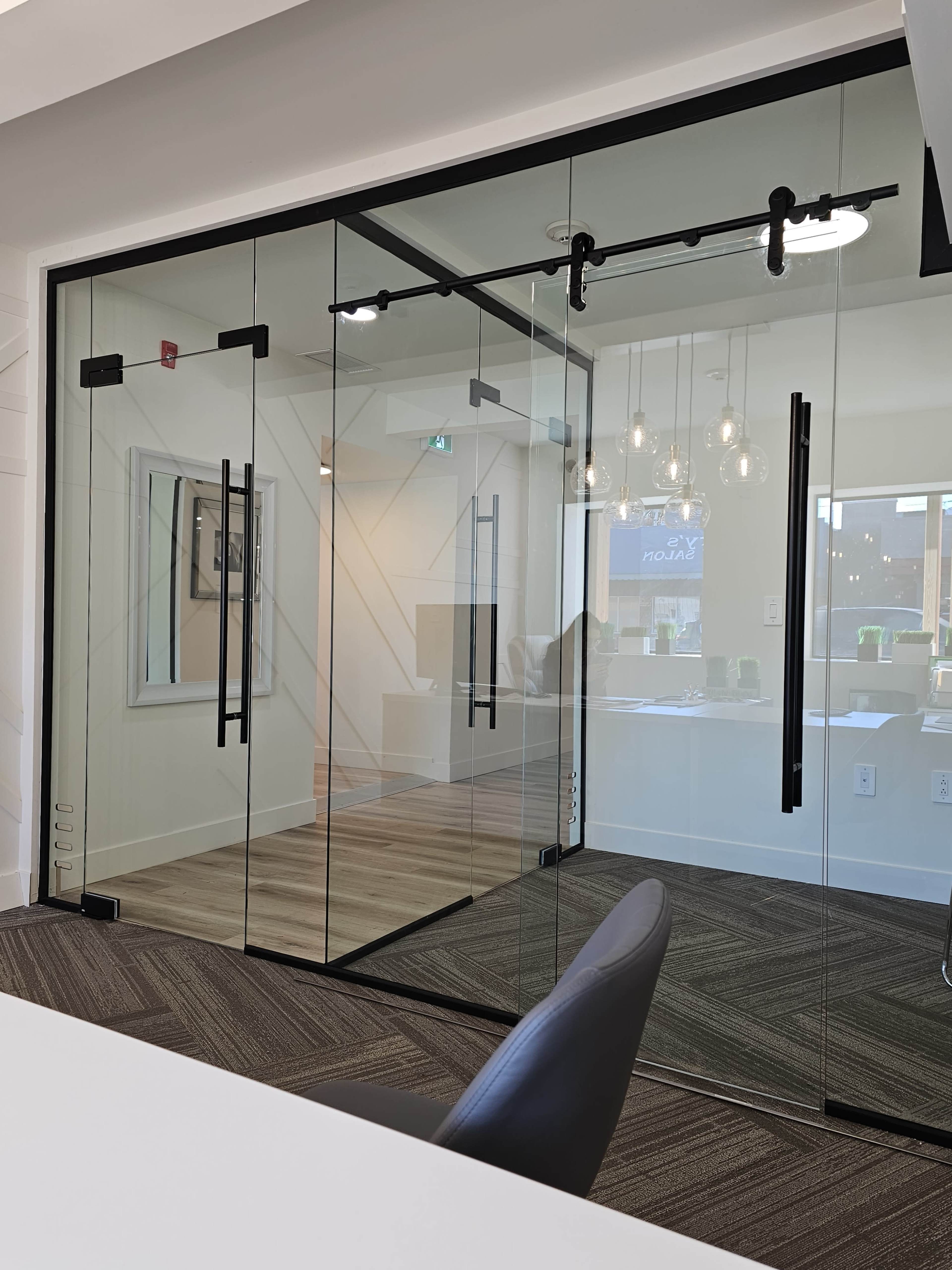 The image shows a modern office space featuring a glass meeting room with black handles and a minimalist design, illuminated by pendant lights.