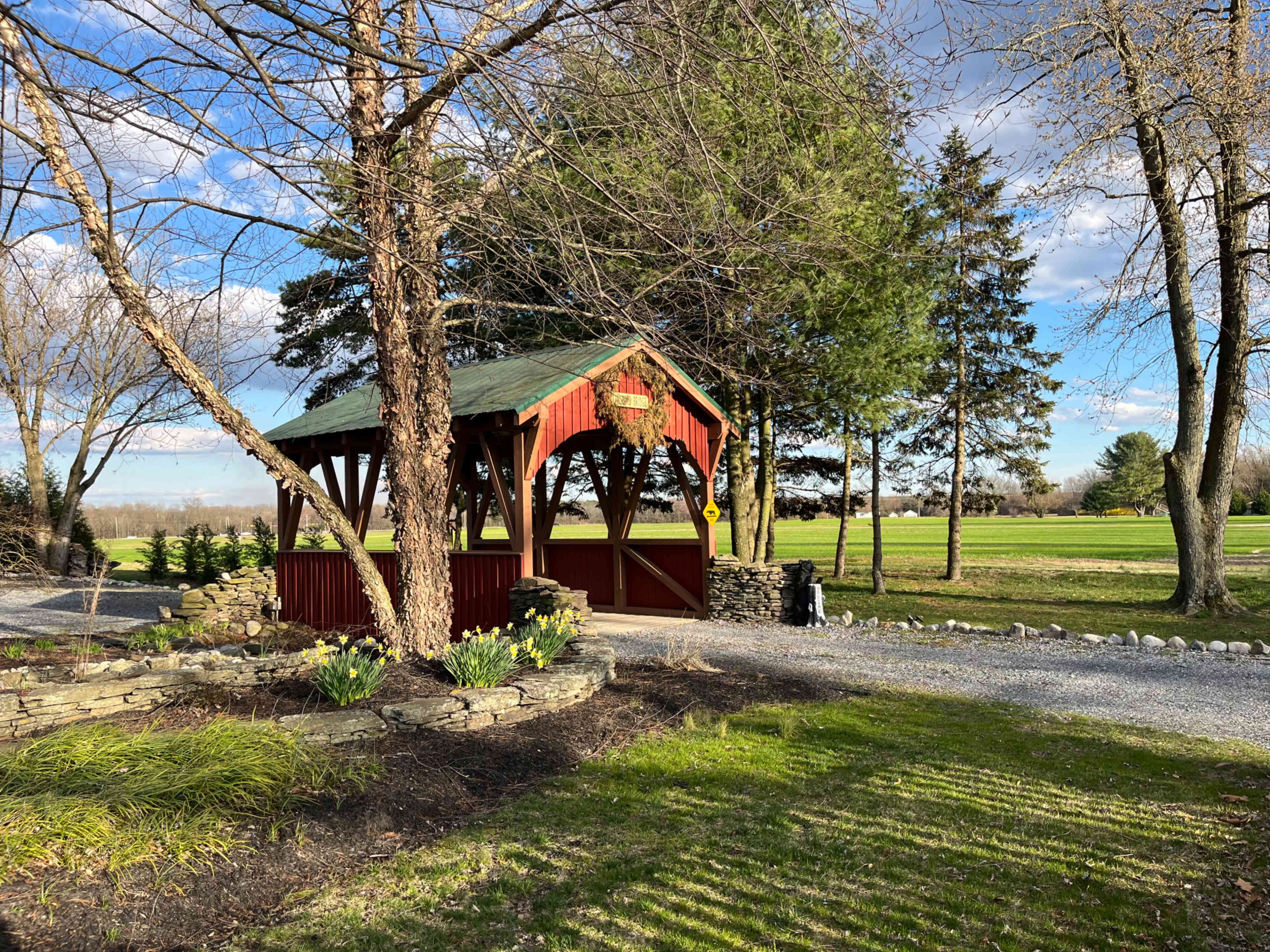 A red-and-green covered bridge is surrounded by trees and a garden with daffodils, set against a backdrop of an open field and blue sky.
