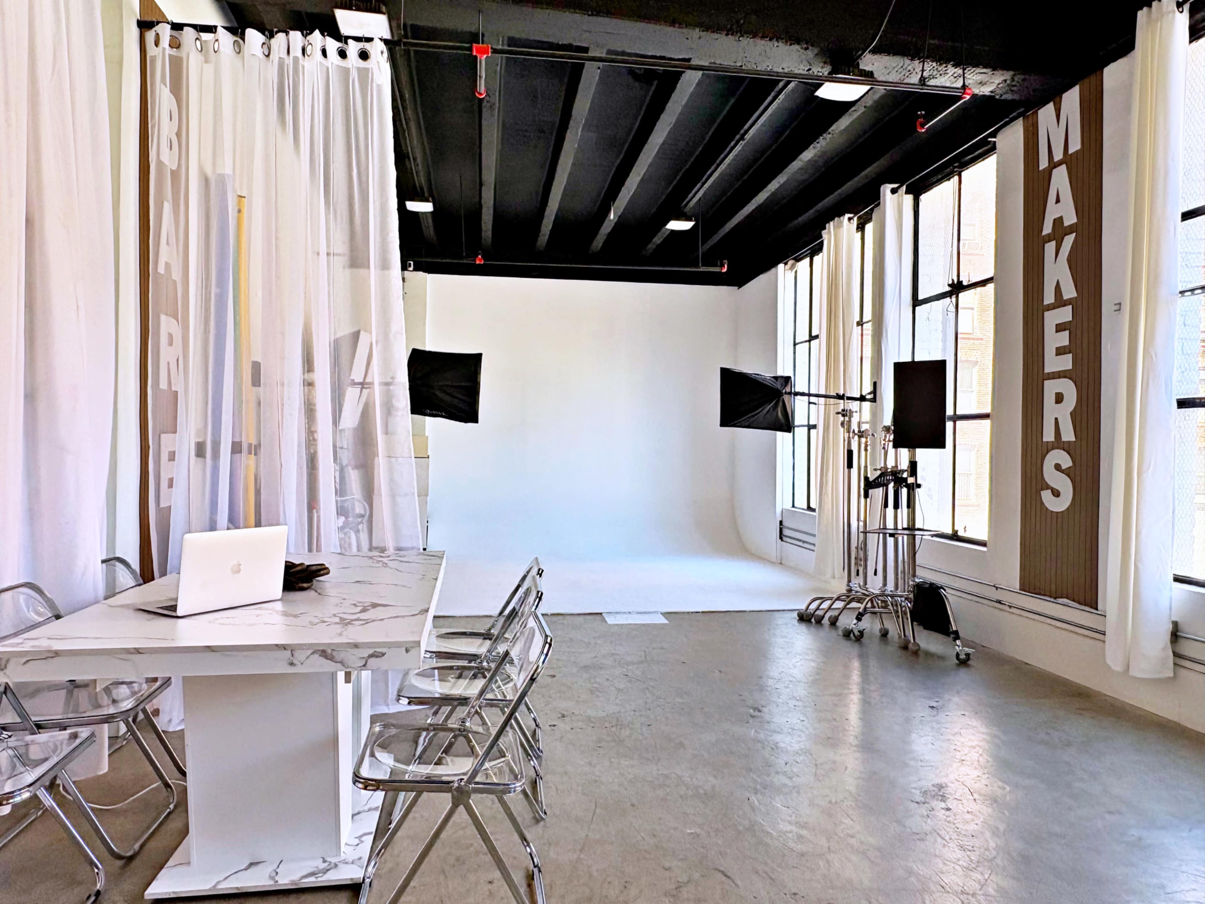 The image shows a bright studio space featuring a white backdrop, photography lights, a modern marble table with chairs, and large windows allowing natural light in.