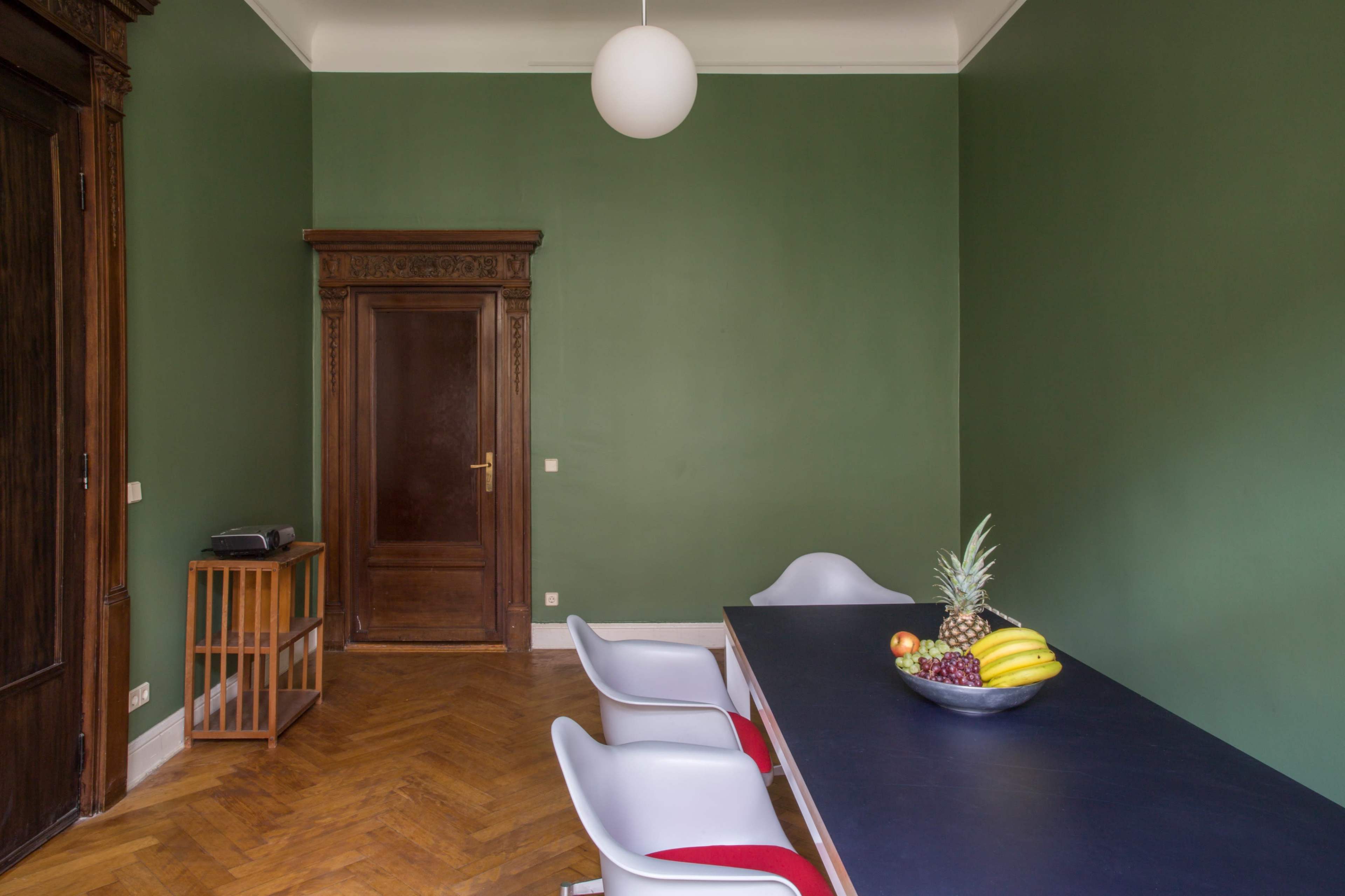 A room with green walls, a wooden door, a wooden cabinet, a table set with a fruit bowl, and three white chairs.