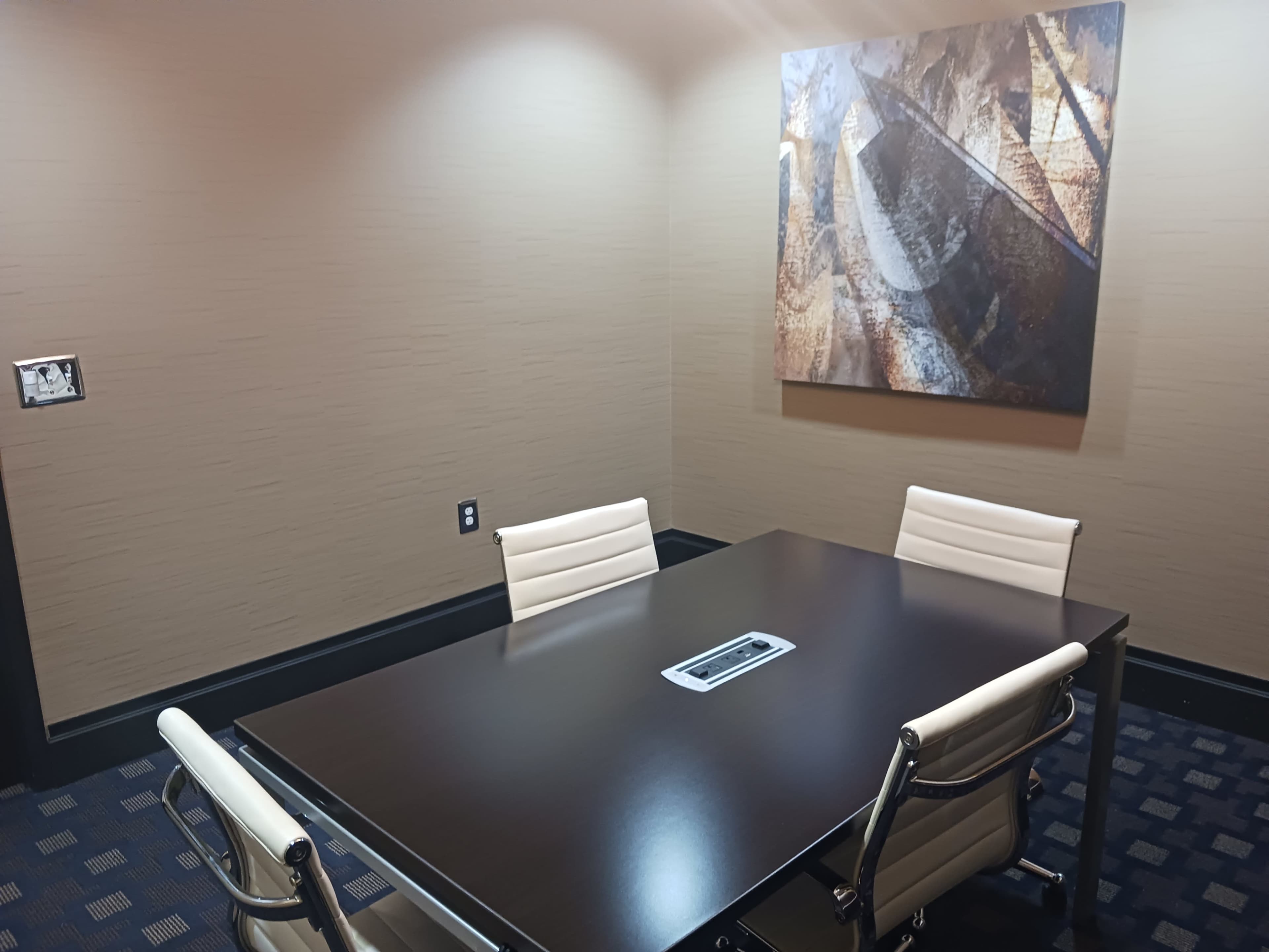 A small conference room with a dark wooden table and four white chairs, accompanied by a large abstract painting on the wall.