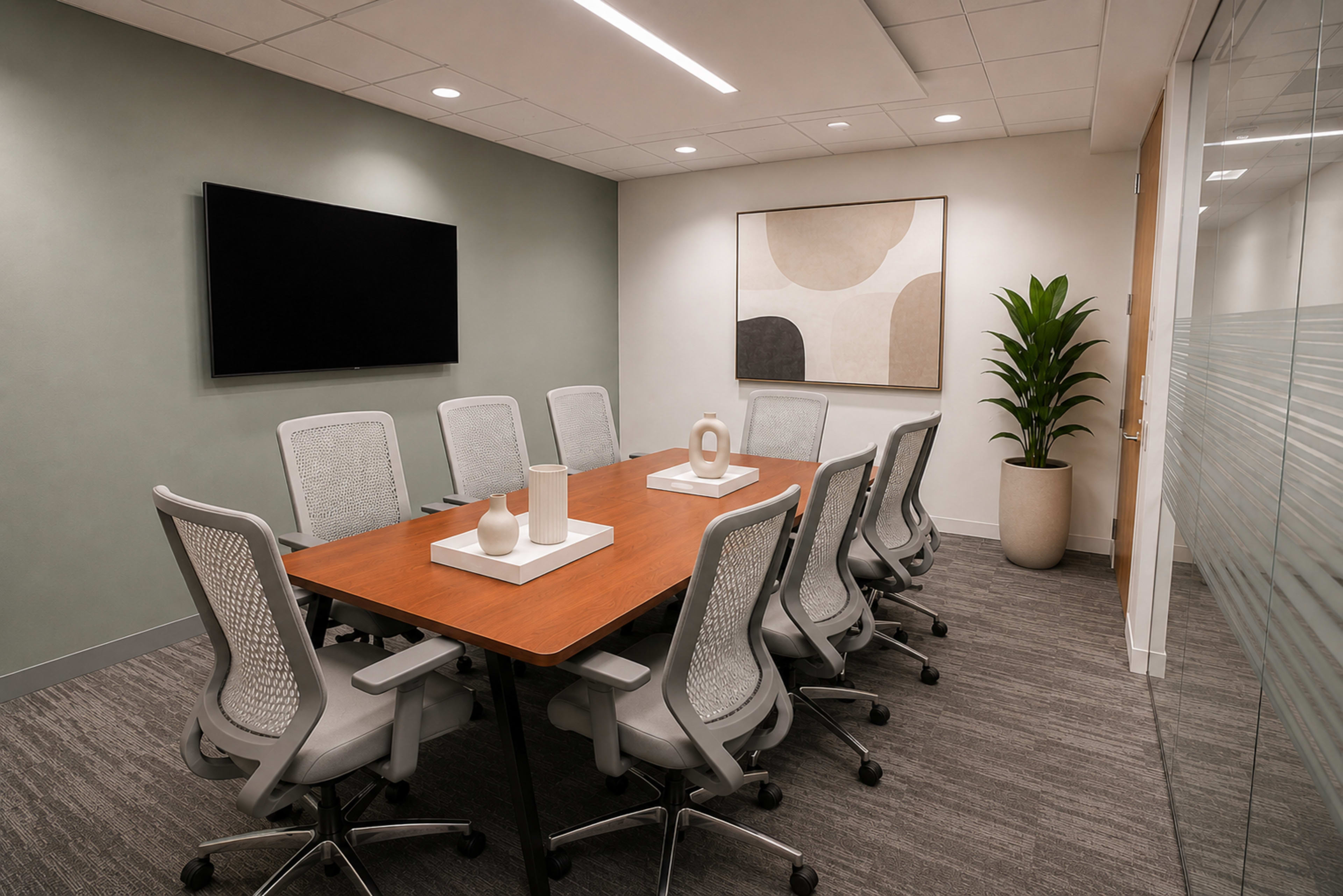 A modern conference room features a large wooden table surrounded by ergonomic chairs, a wall-mounted TV, and decorative elements on the table along with a potted plant in the corner.