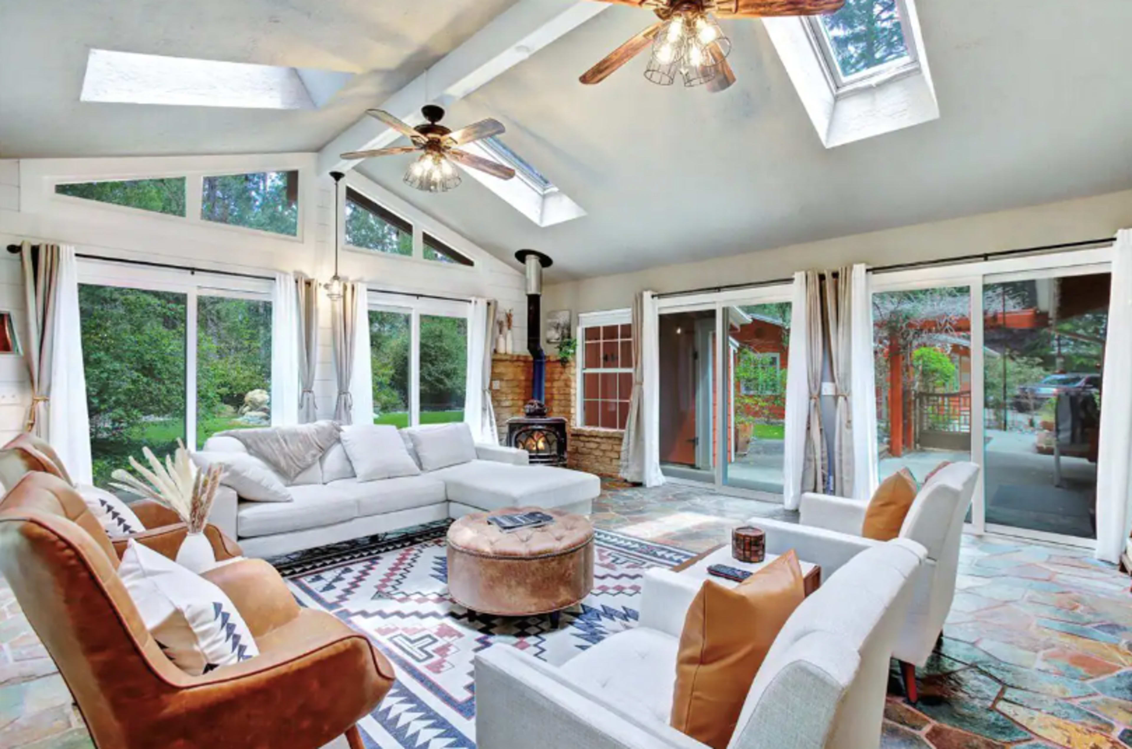 A spacious living room with a sectional sofa, a round coffee table, large windows, and skylights, featuring a stone floor and a wood-burning stove.