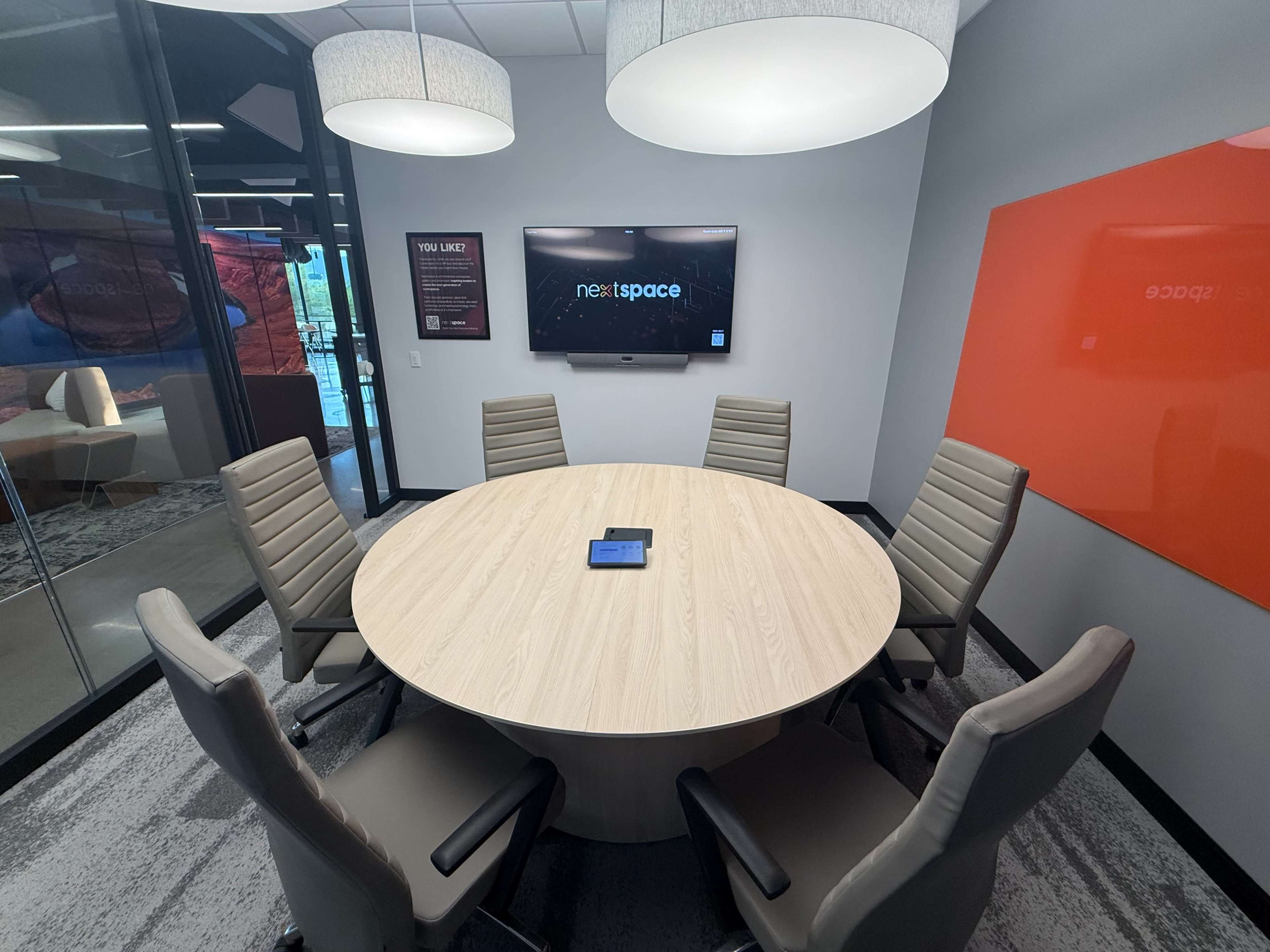 A modern conference room features a round wooden table surrounded by six high-backed chairs, with a large screen mounted on the wall displaying the "nextspace" logo.