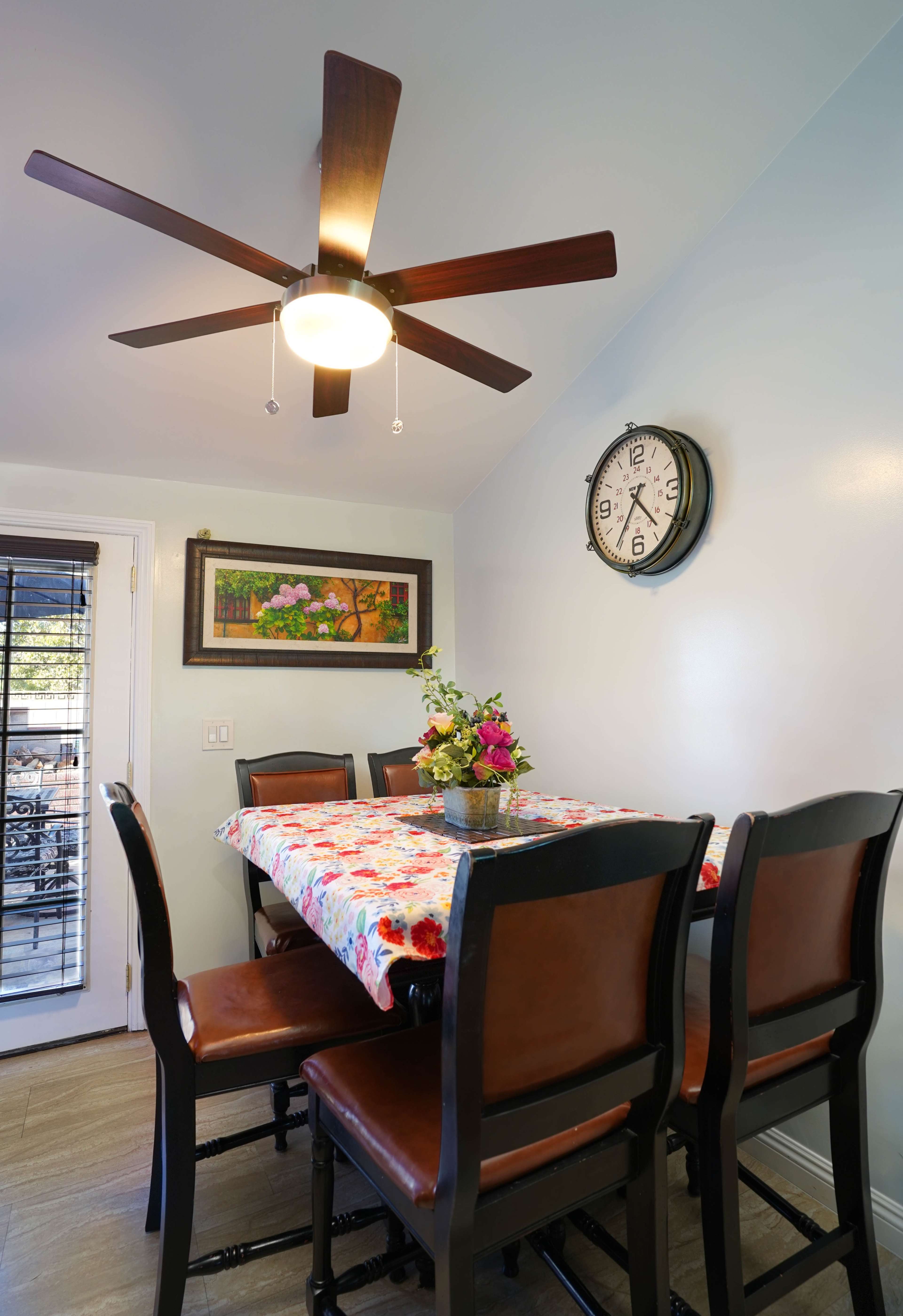 A dining area features a table covered with a floral tablecloth surrounded by high-backed chairs, with a ceiling fan and wall clock above.
