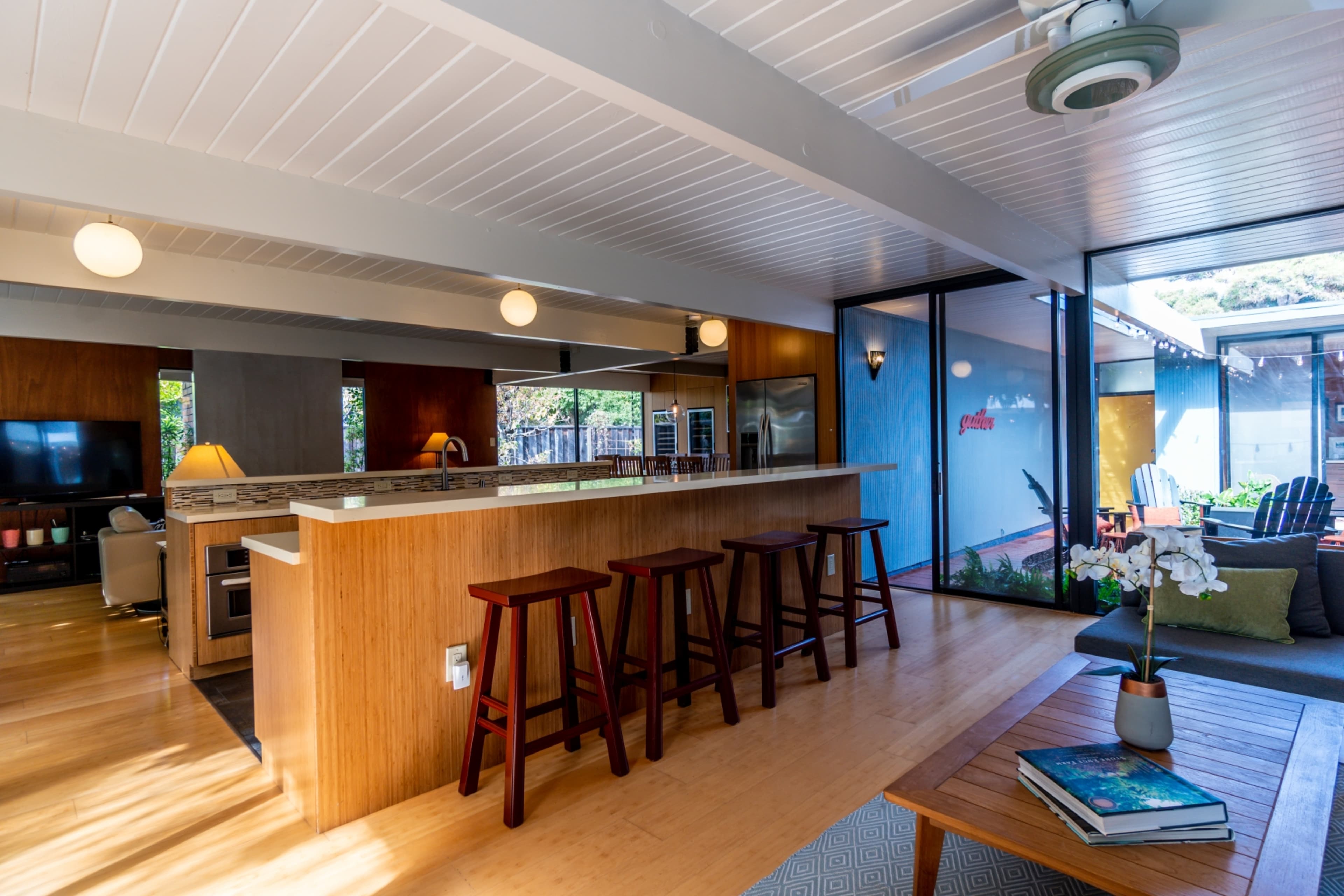 The image shows a modern open-concept kitchen and living area with wooden cabinetry, a central island, and large glass windows.