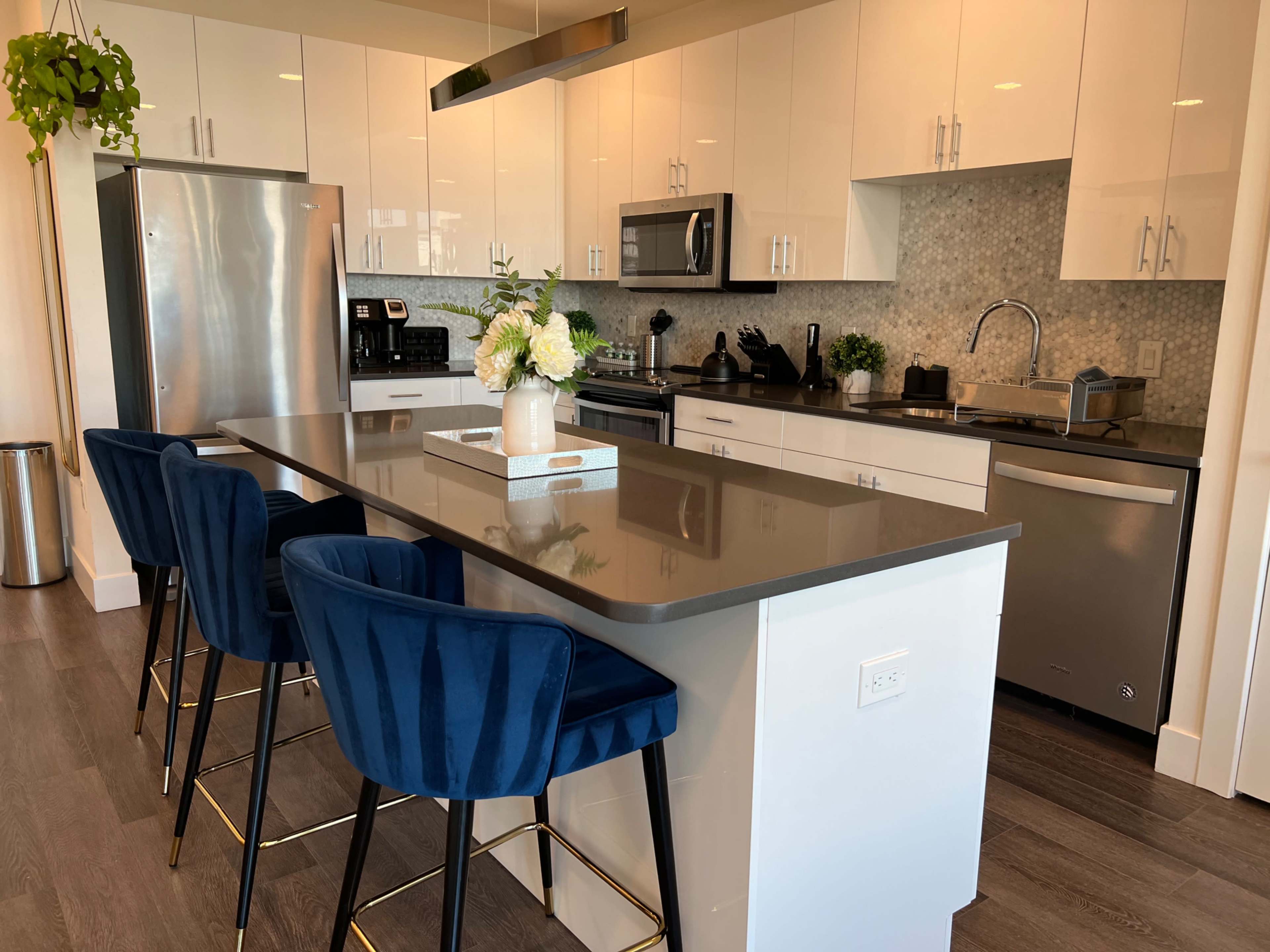 A modern kitchen with a large island featuring four blue stools, stainless steel appliances, and white cabinetry.