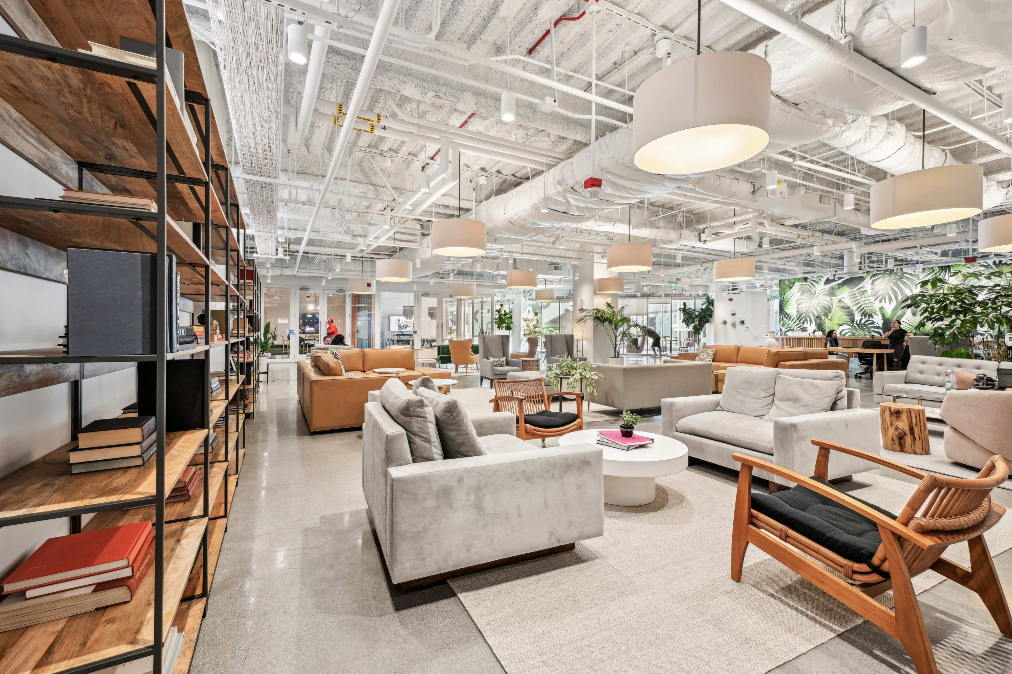 The image shows a spacious modern office interior featuring a lounge area with various seating arrangements, large windows, and greenery.