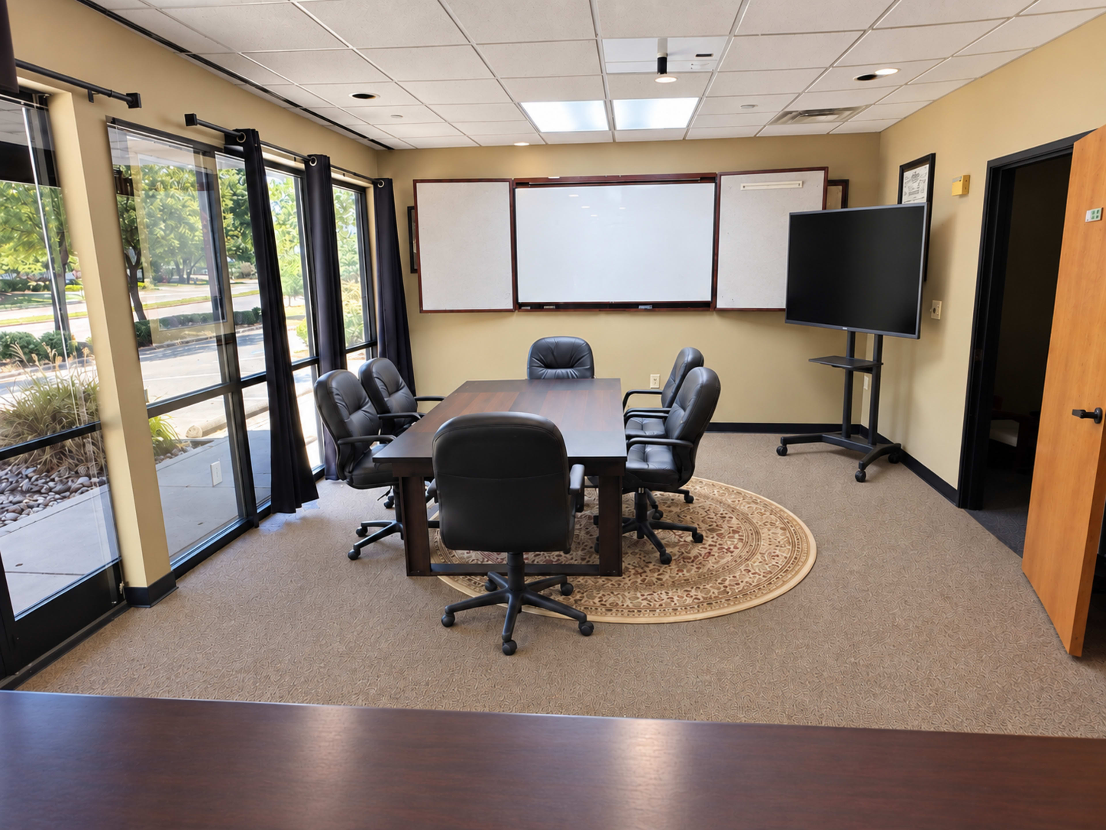 The image shows a conference room featuring a large table surrounded by six black chairs, a whiteboard on the wall, a screen on a stand, and large windows allowing natural light to enter.