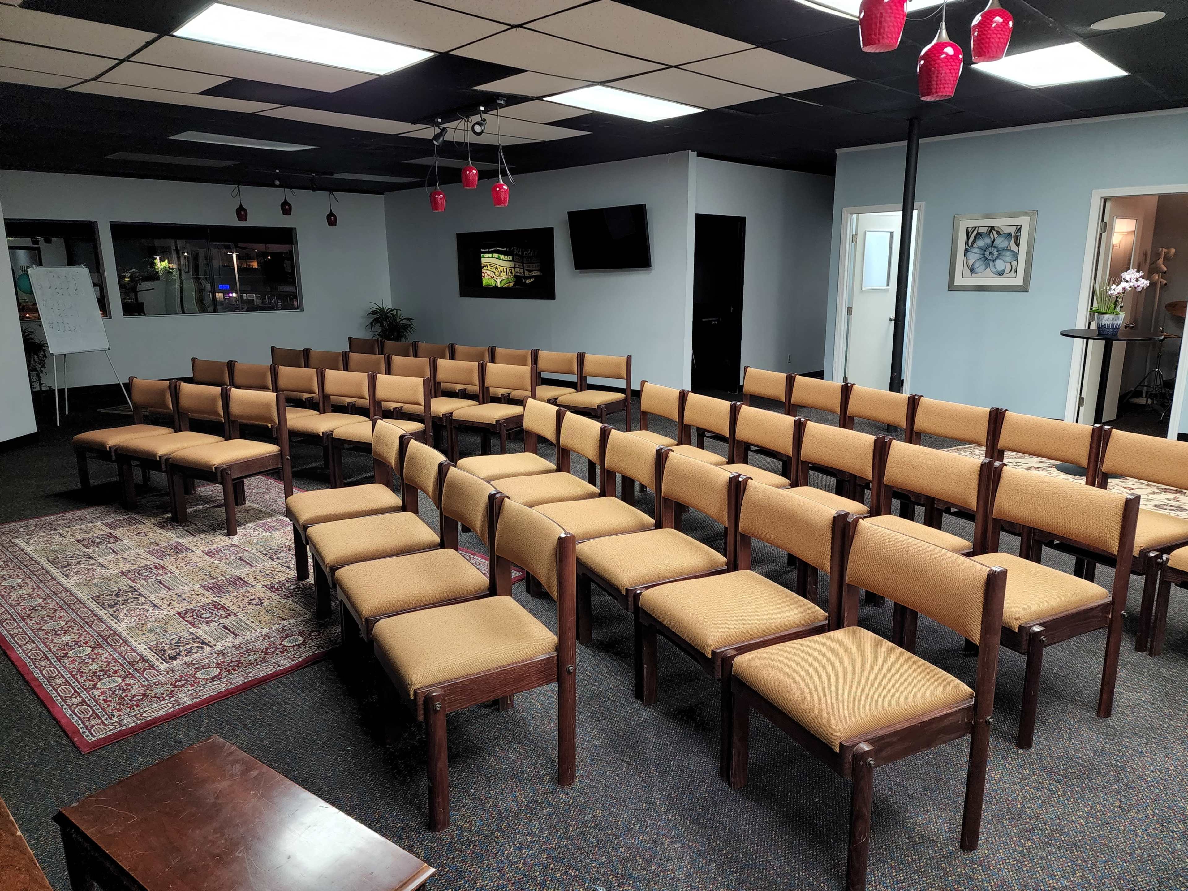 A room arranged with rows of wooden chairs featuring upholstered seats, set up in a semi-circle around a central space, with a patterned rug on the floor and overhead lighting fixtures.