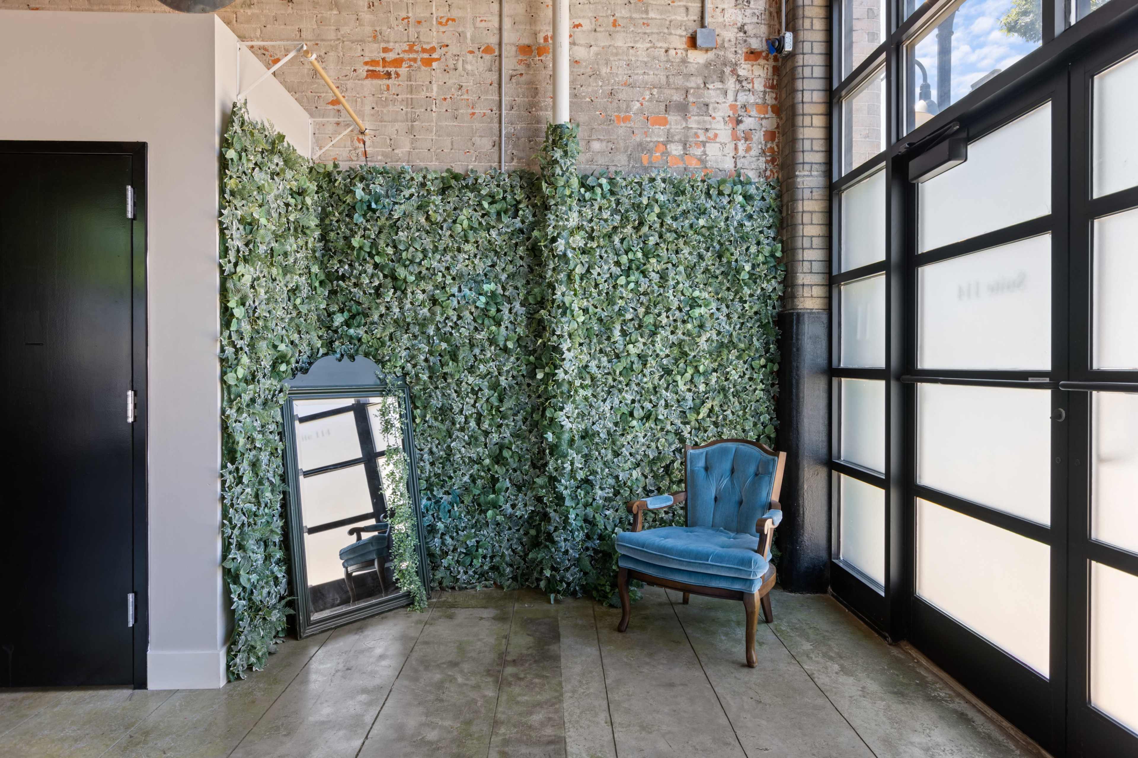 The image depicts a corner of a room with a large mirror and a blue upholstered chair next to a wall covered in greenery, alongside a glass door.