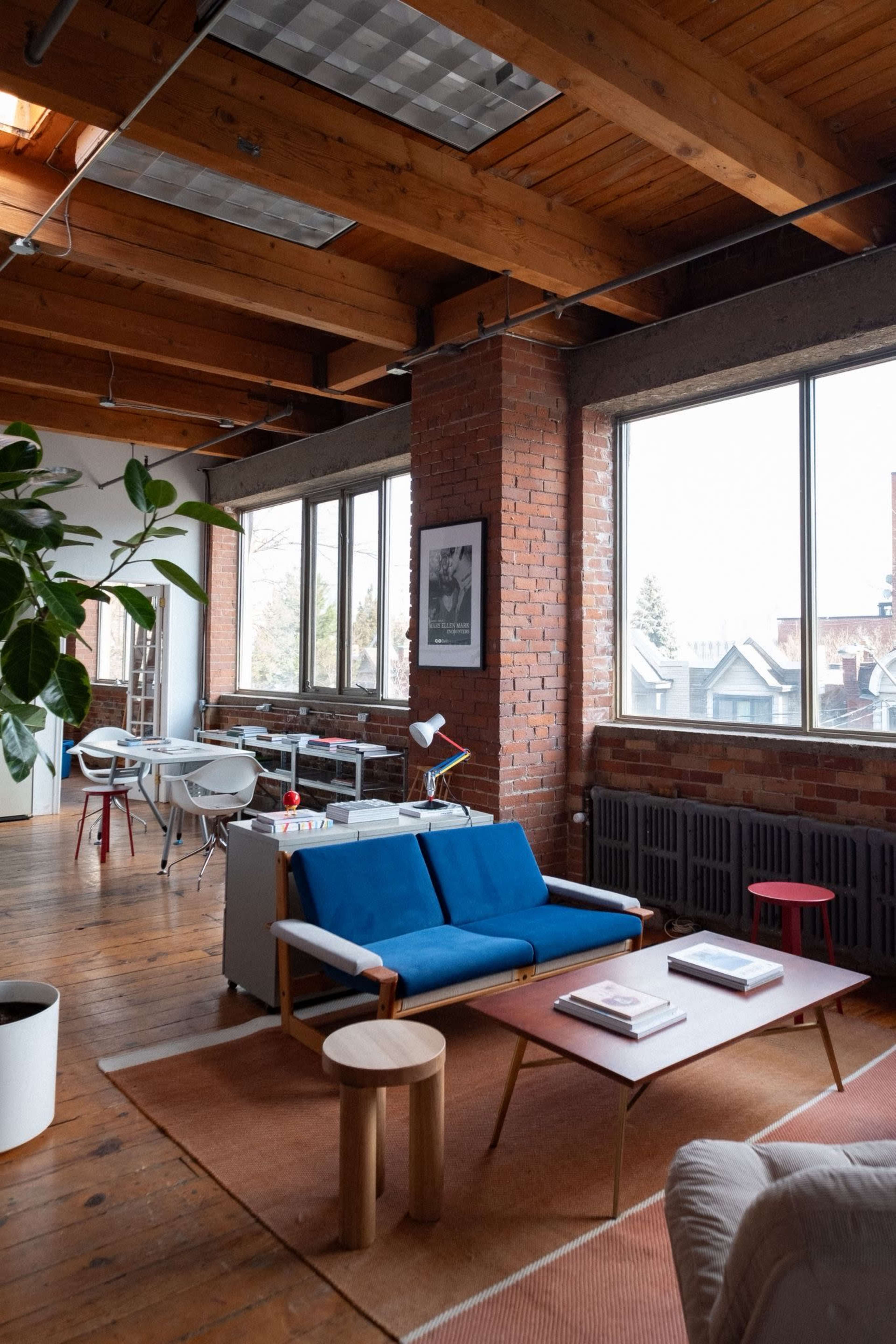The image shows a modern office interior featuring wooden beams, a brick wall, large windows, and a mix of contemporary furniture including a blue sofa and wooden tables.