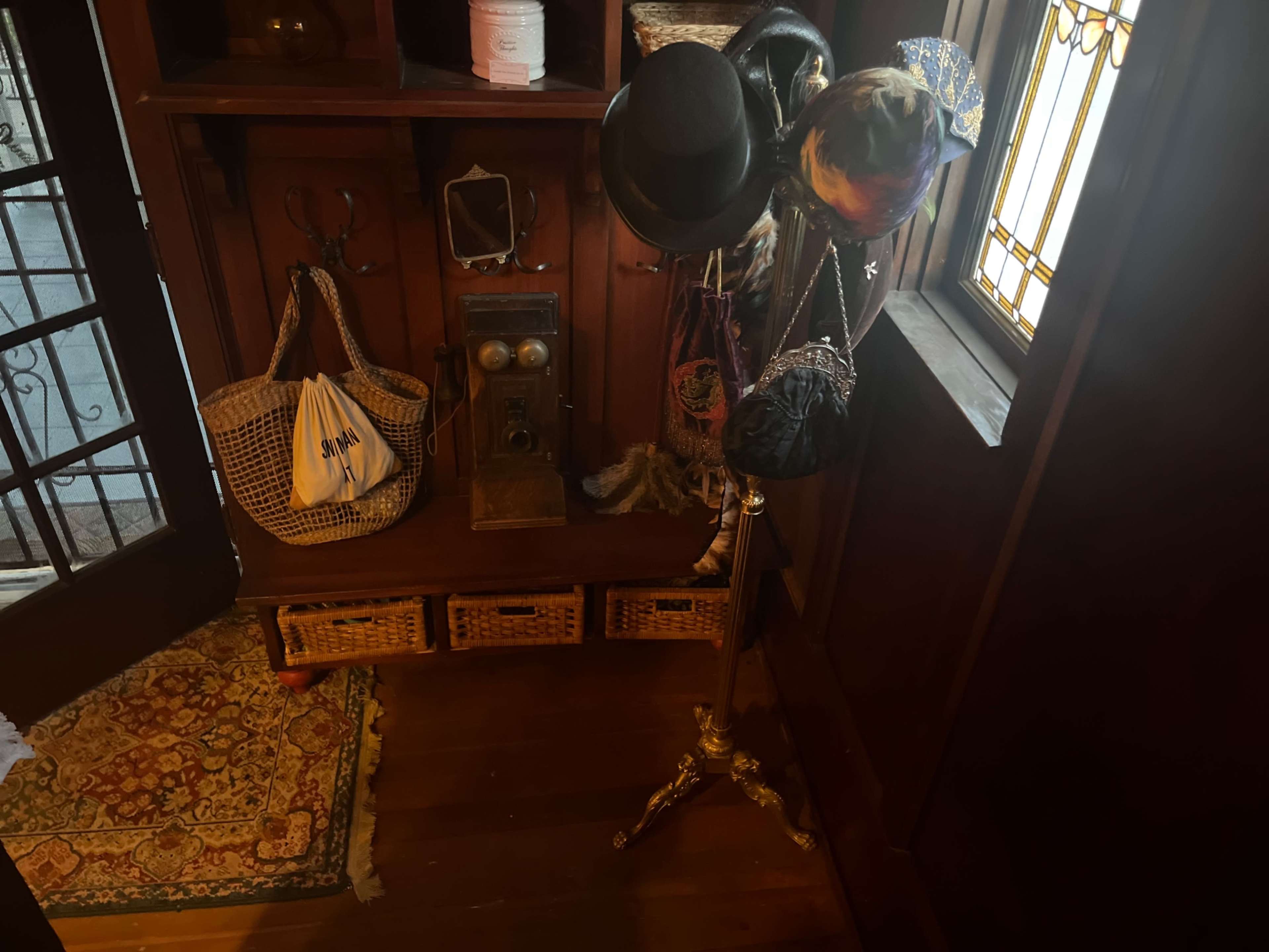 The image shows a wooden interior featuring a coat rack with hats, a vintage phone, and woven baskets.