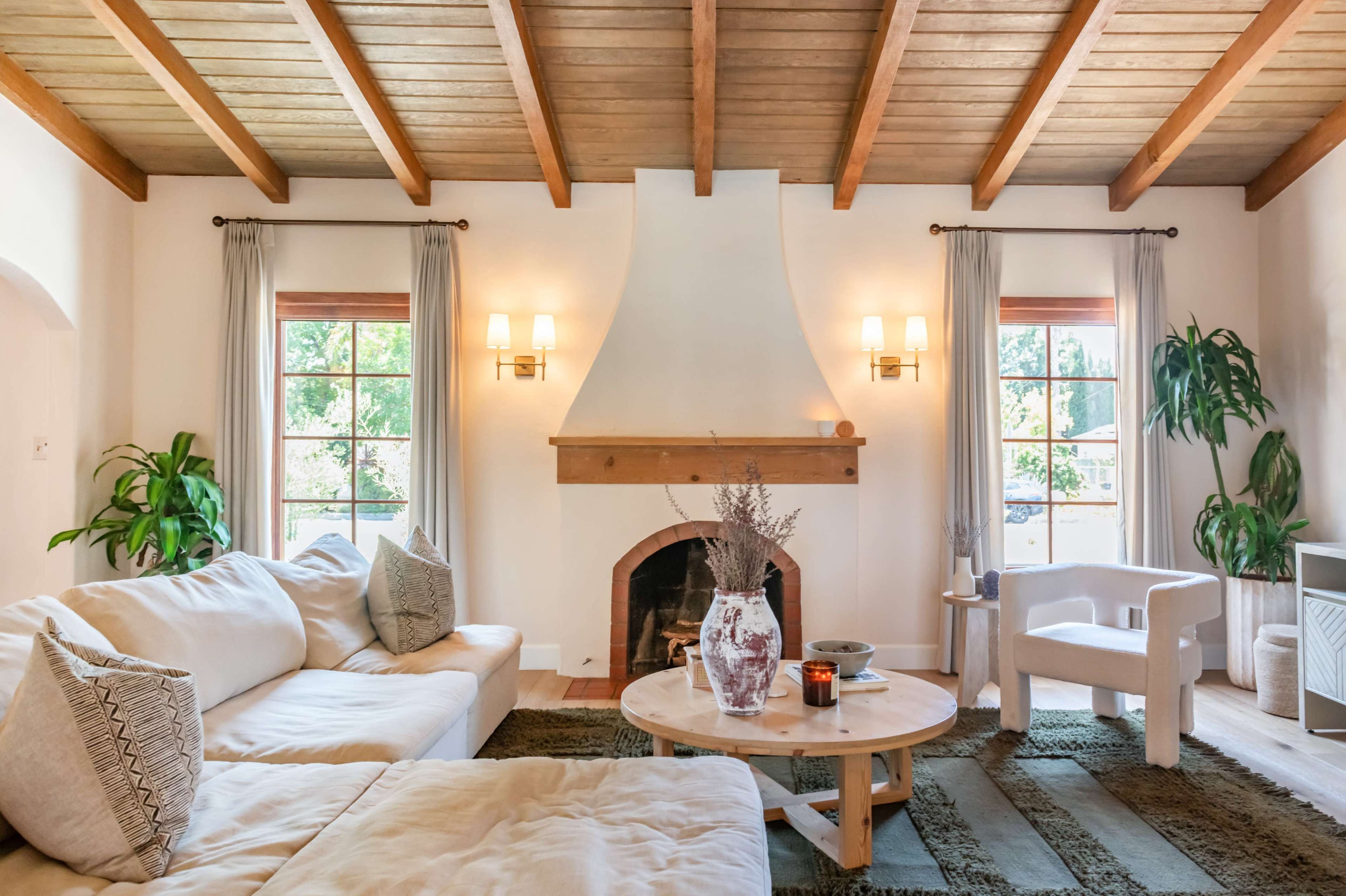 The image shows a cozy living room with a beige sofa, a round coffee table, a fireplace, and large windows framed by light curtains, complemented by indoor plants and wood beams on the ceiling.