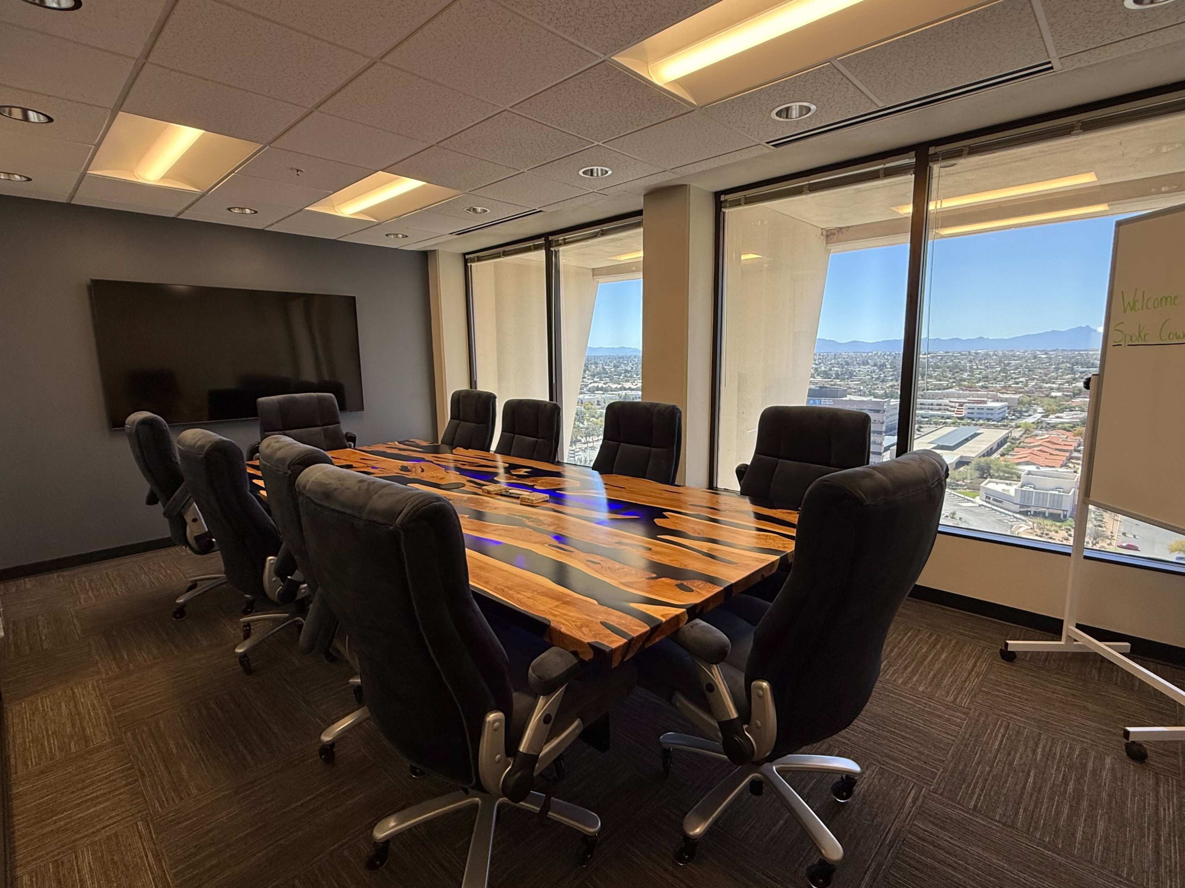 A large conference room features a wooden table with blue accents, surrounded by ergonomic chairs and large windows displaying a city view.