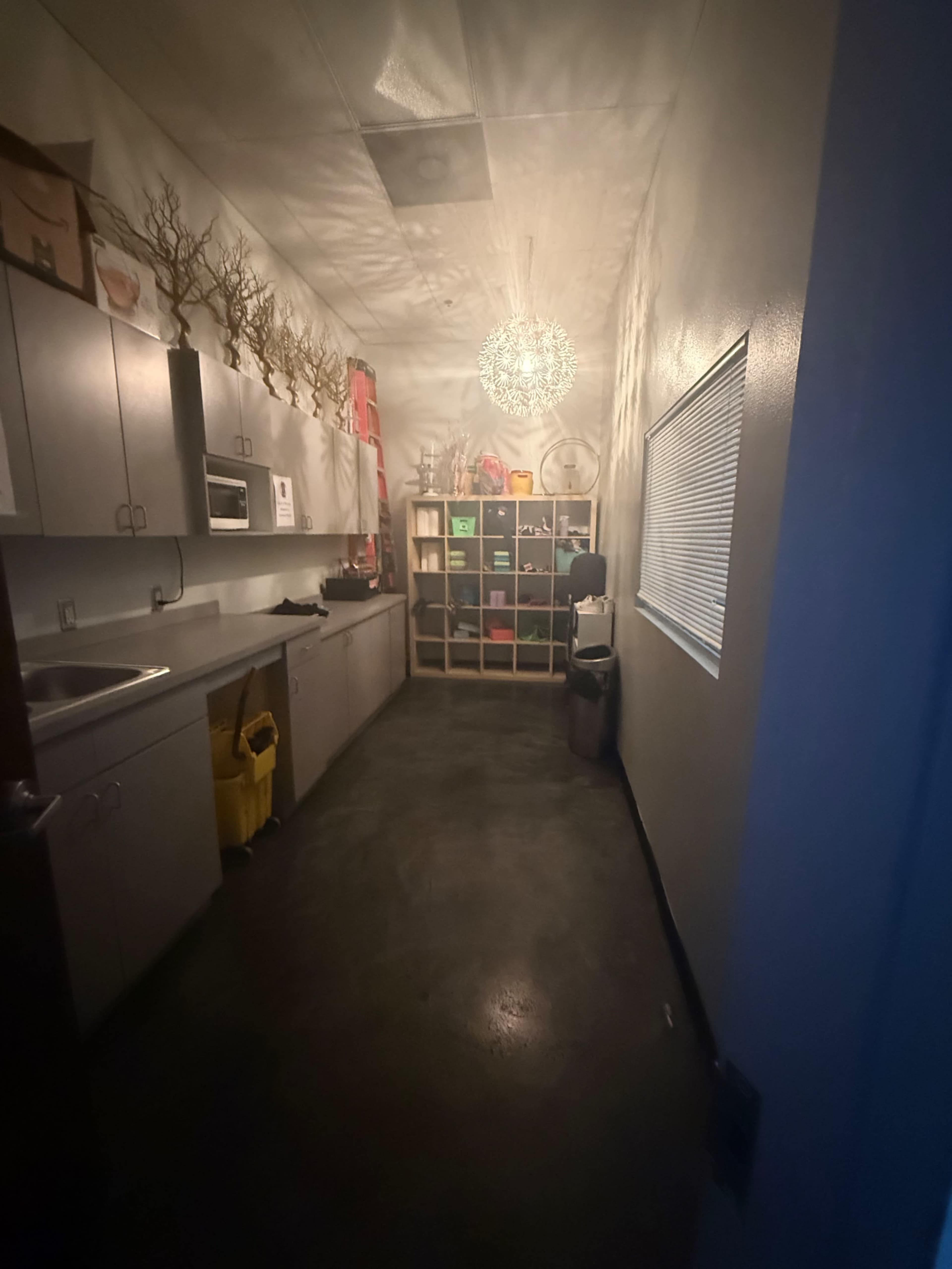 The image depicts a dimly lit kitchen or breakroom with cabinets, a countertop, and a decorative shelf filled with colorful storage bins.