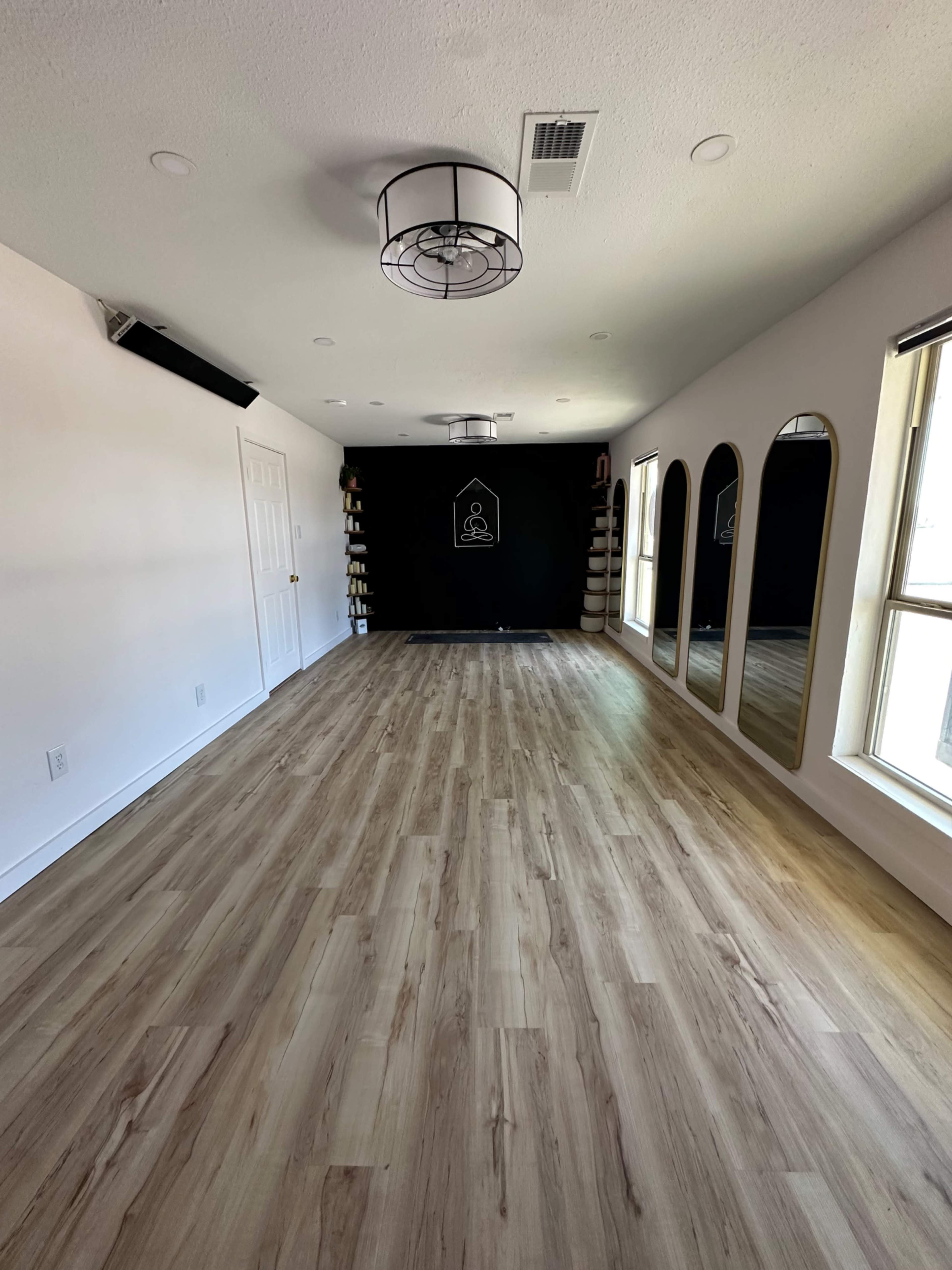 The image shows a spacious, well-lit room with wooden flooring, a dark accent wall featuring a symbol, and three large mirrors framed on one side.