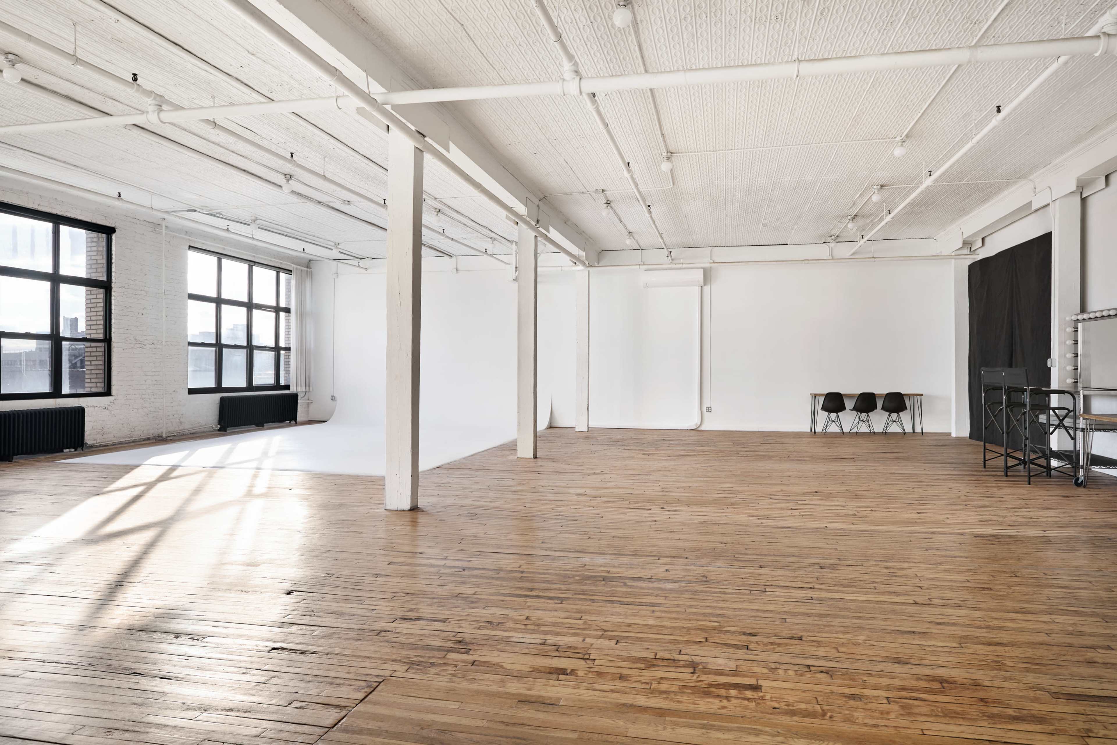 The image shows a spacious, empty studio with hardwood floors, white walls, and large windows letting in natural light.