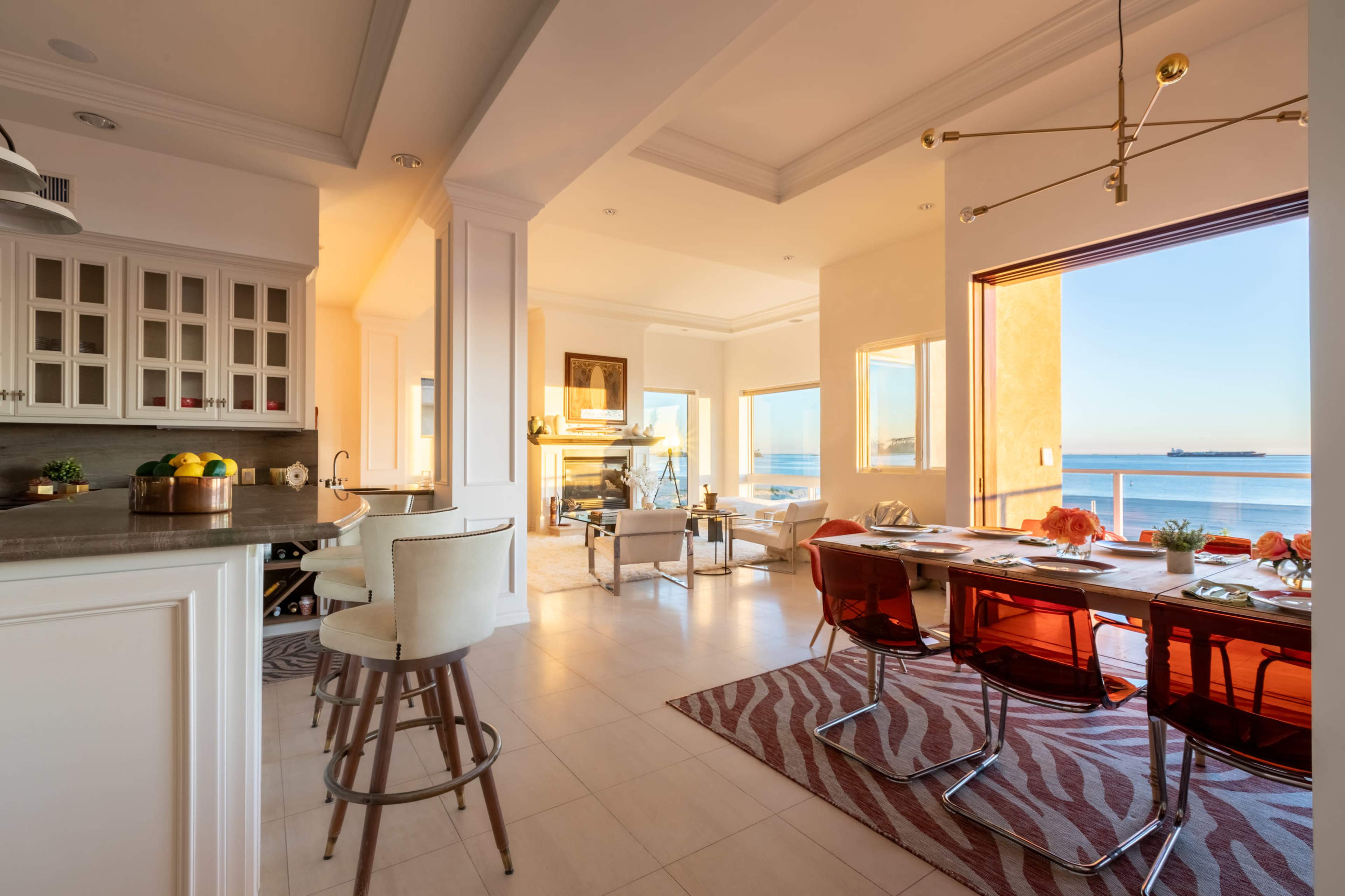 The image shows a modern open-concept living space featuring a kitchen, dining area, and large windows with a view of the ocean.