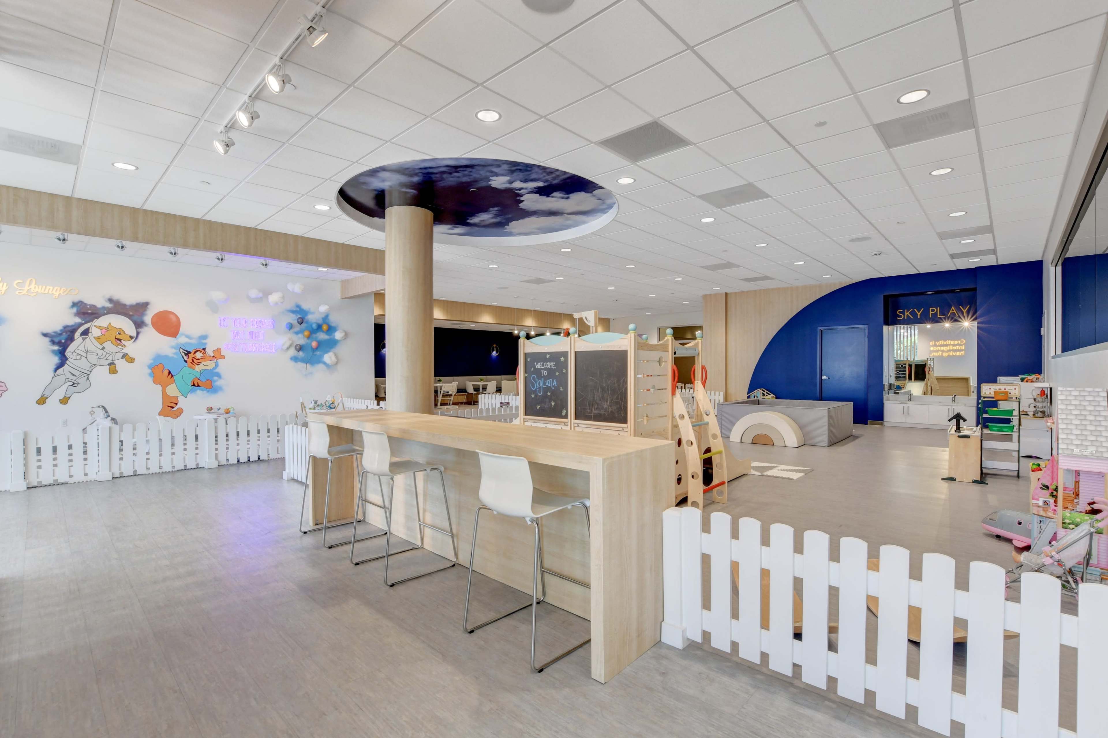 A brightly lit children's play area with a bar-style seating section and various play structures enclosed by white picket fencing.