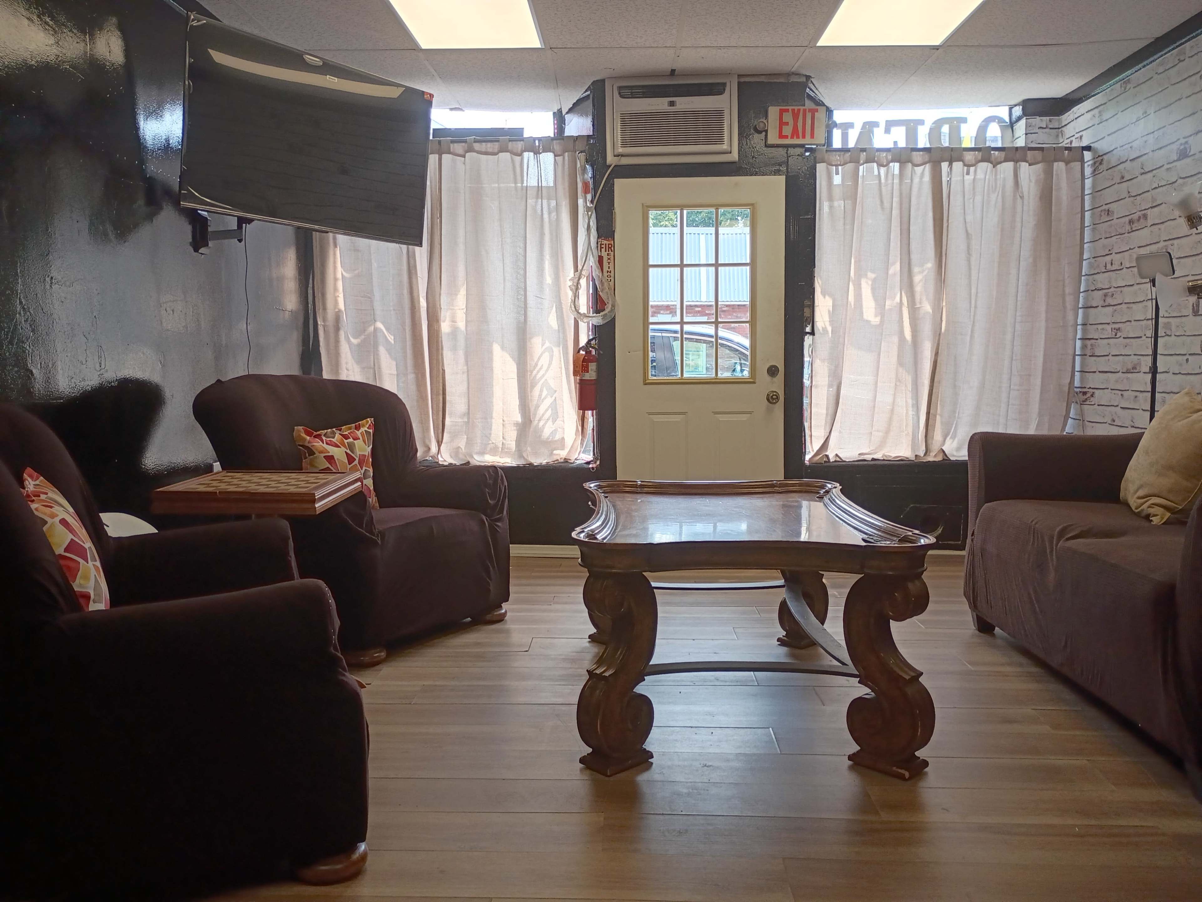 The image shows a cozy waiting area with two dark armchairs, a wooden coffee table, and a door with sheer curtains in front of a window.