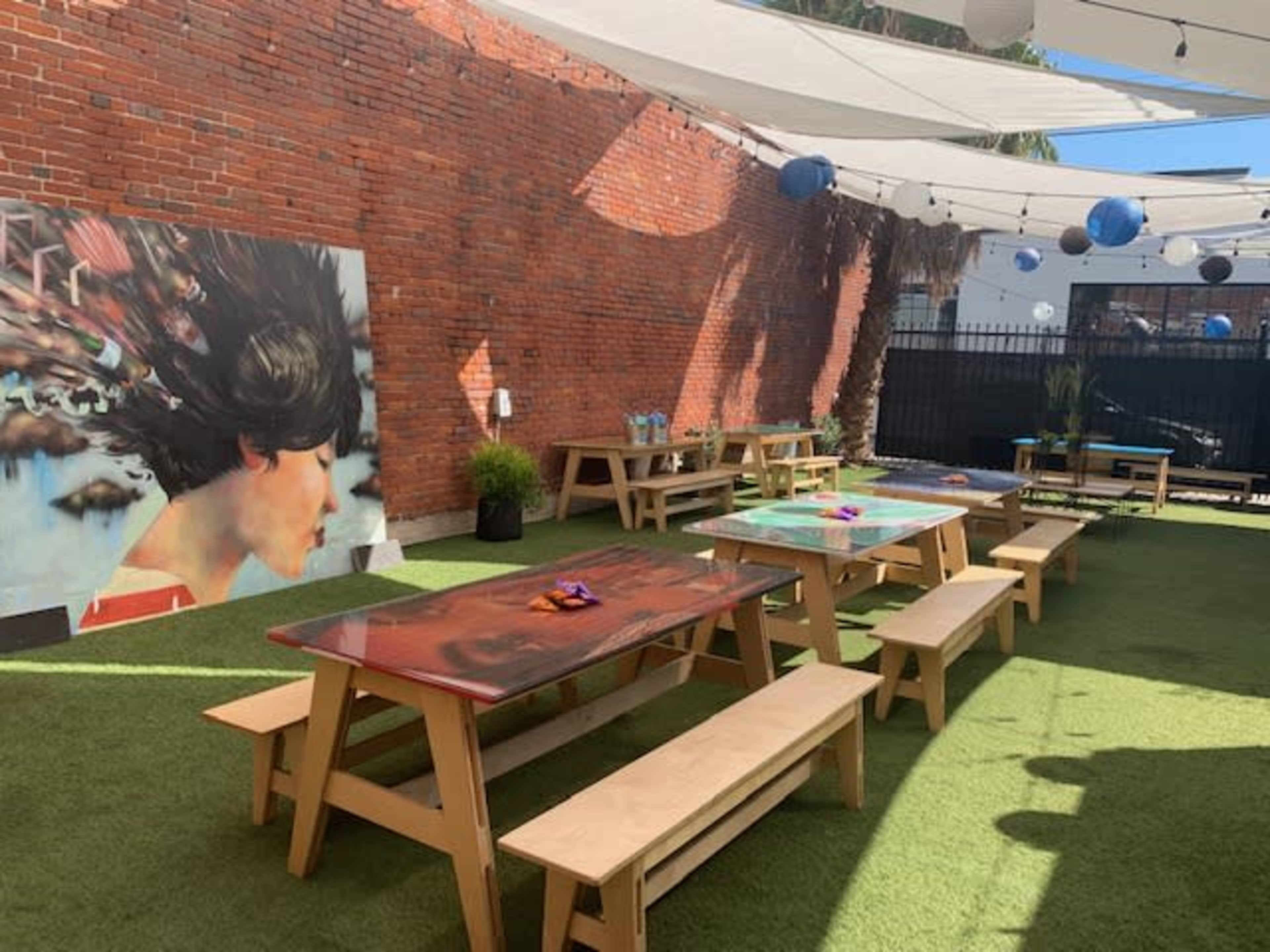 The outdoor space features wooden tables and benches on green grass, with a brick wall adorned by a colorful mural and decorative hanging lanterns.