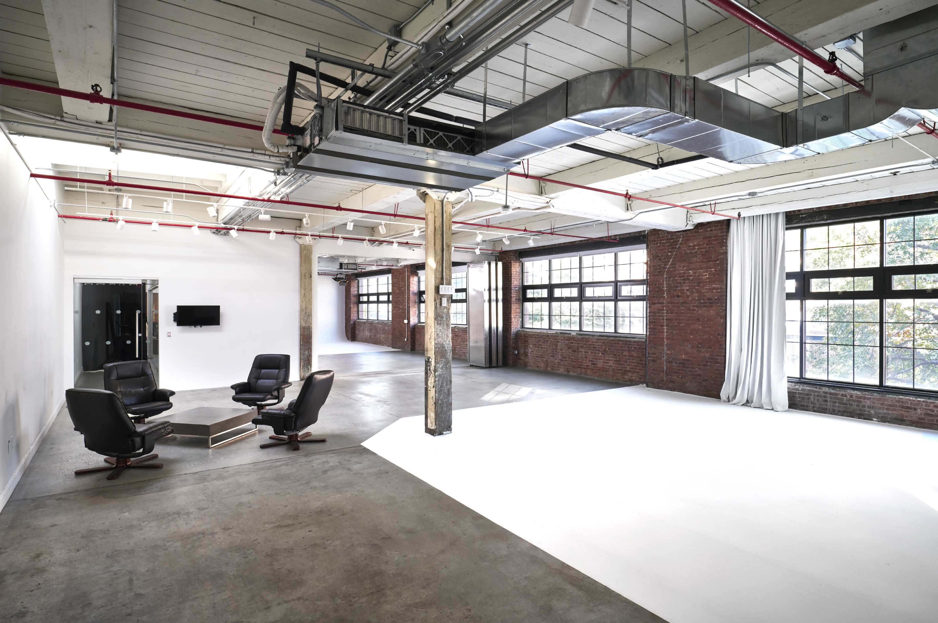 An industrial-style interior space featuring four black chairs arranged in a circle, large windows allowing natural light, and exposed brick and concrete elements.