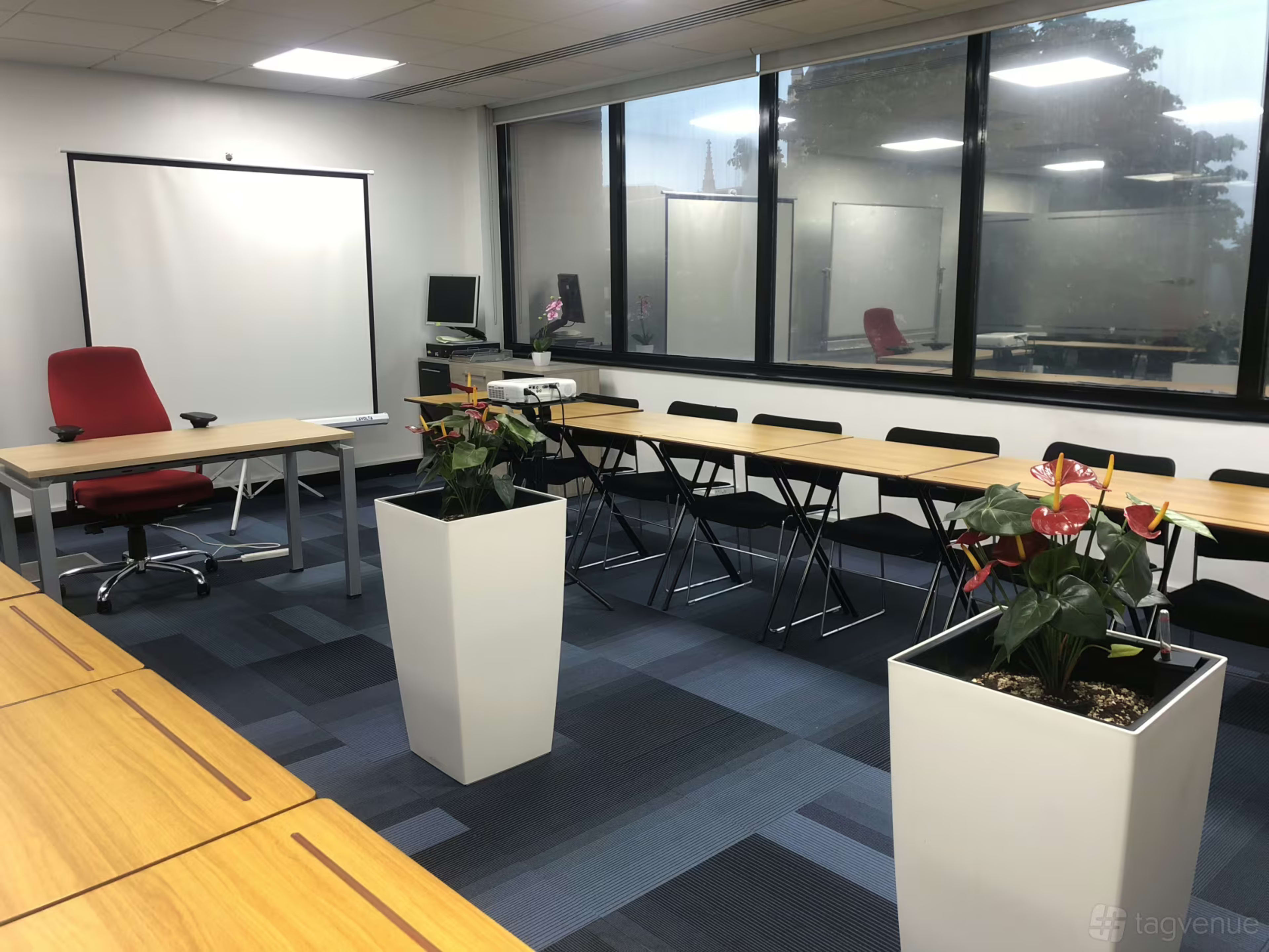 The image shows a modern office meeting room with a large table, chairs, a projector screen, and potted plants.