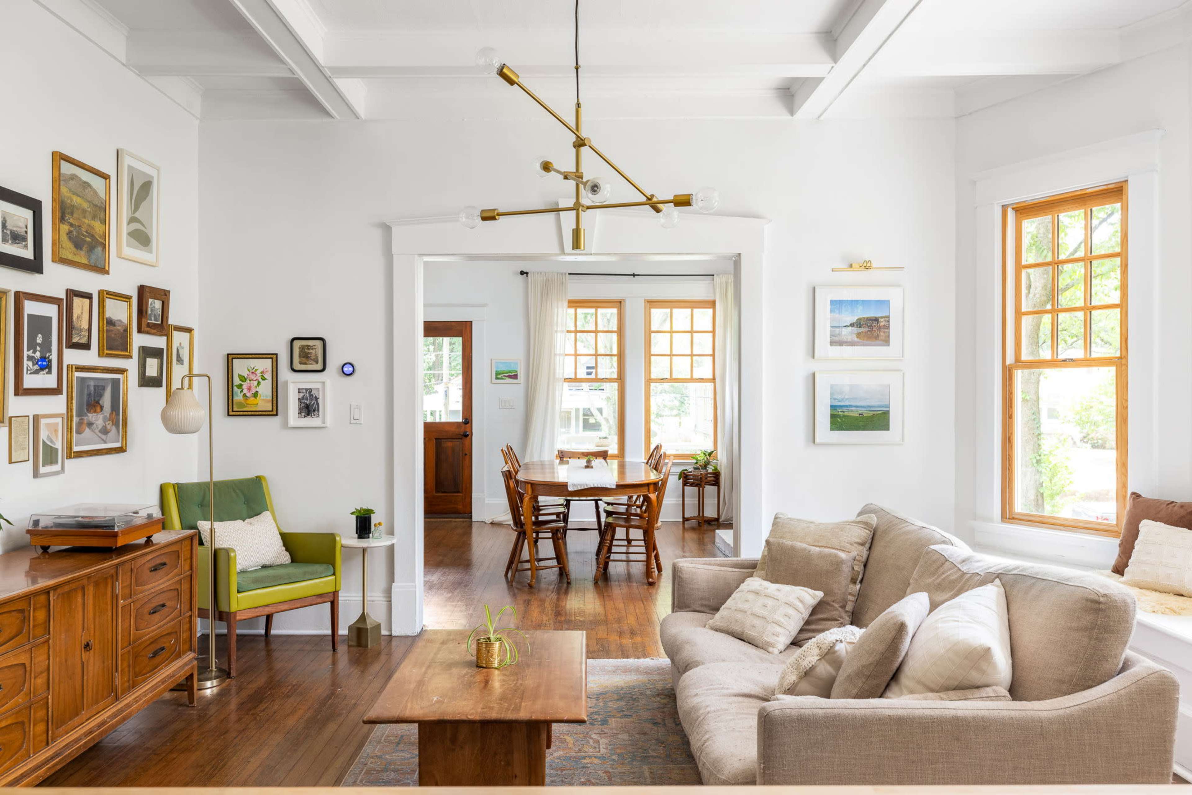A bright living room features a beige sofa, wooden furniture, a green accent chair, and a dining area visible through an archway.