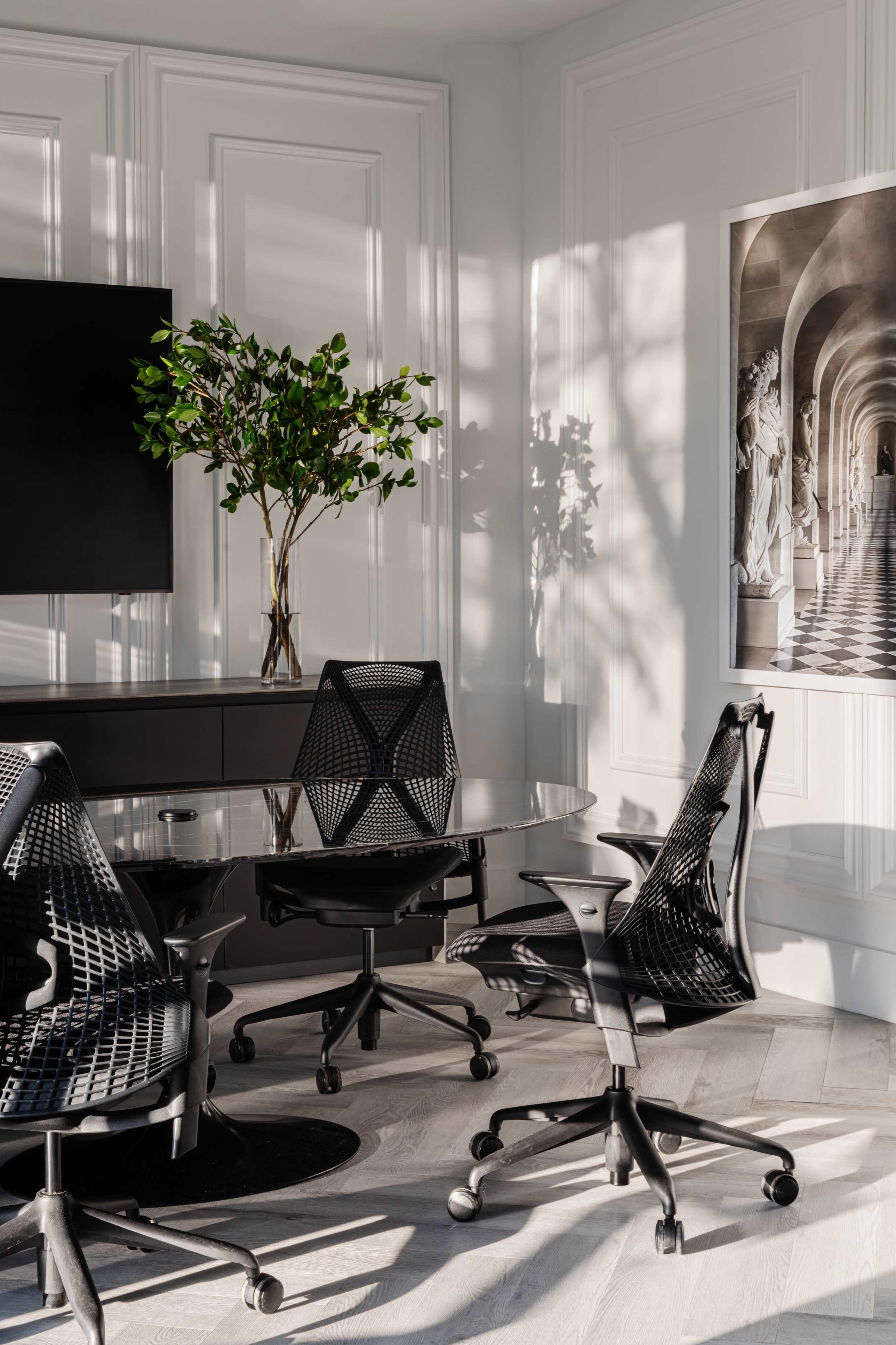 A modern conference room features a glass tabletop with four ergonomic black chairs, illuminated by natural light filtering through the windows.