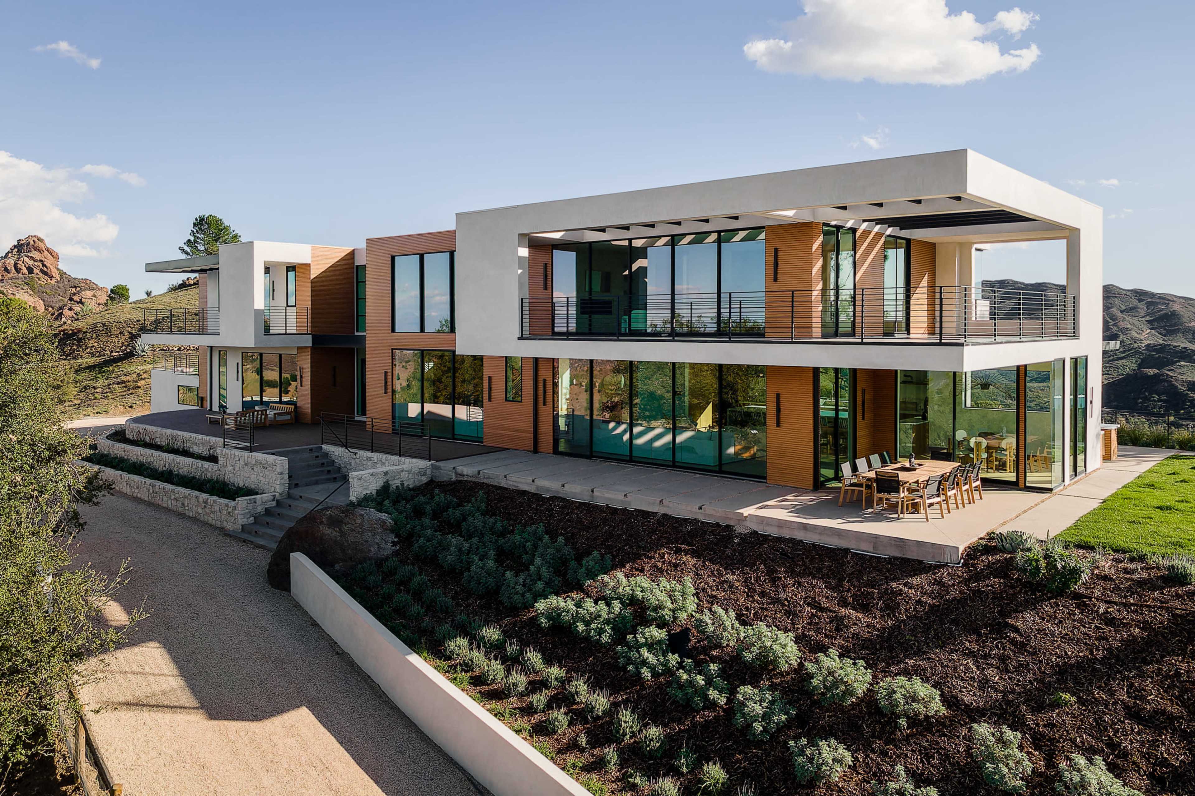 The image shows a modern two-story house with large windows, stone and wooden accents, and a landscaped yard, set against a backdrop of rocky hills.