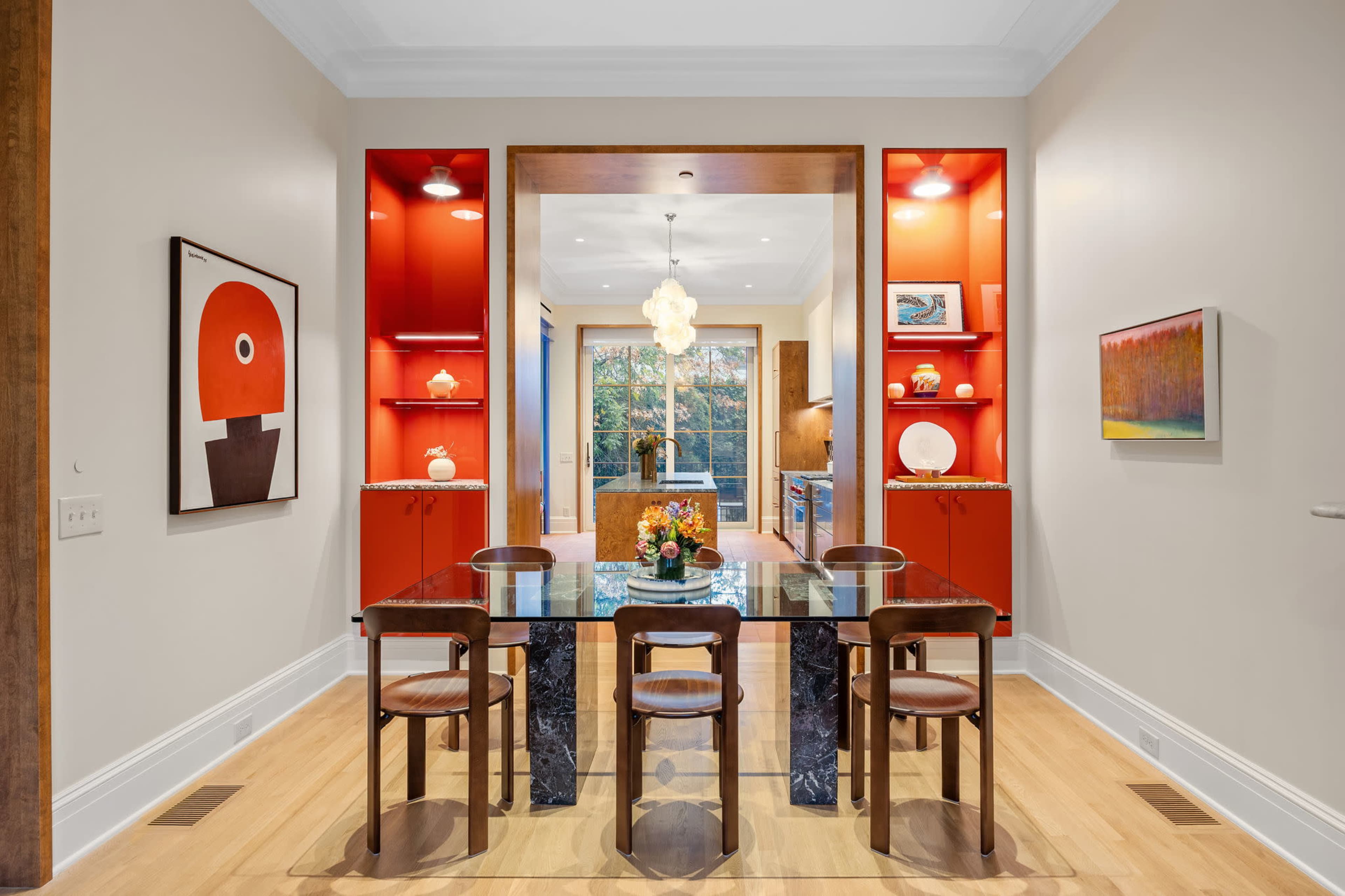 A dining area features a glass table surrounded by four wooden chairs, with red shelves and a colorful artwork on the walls.