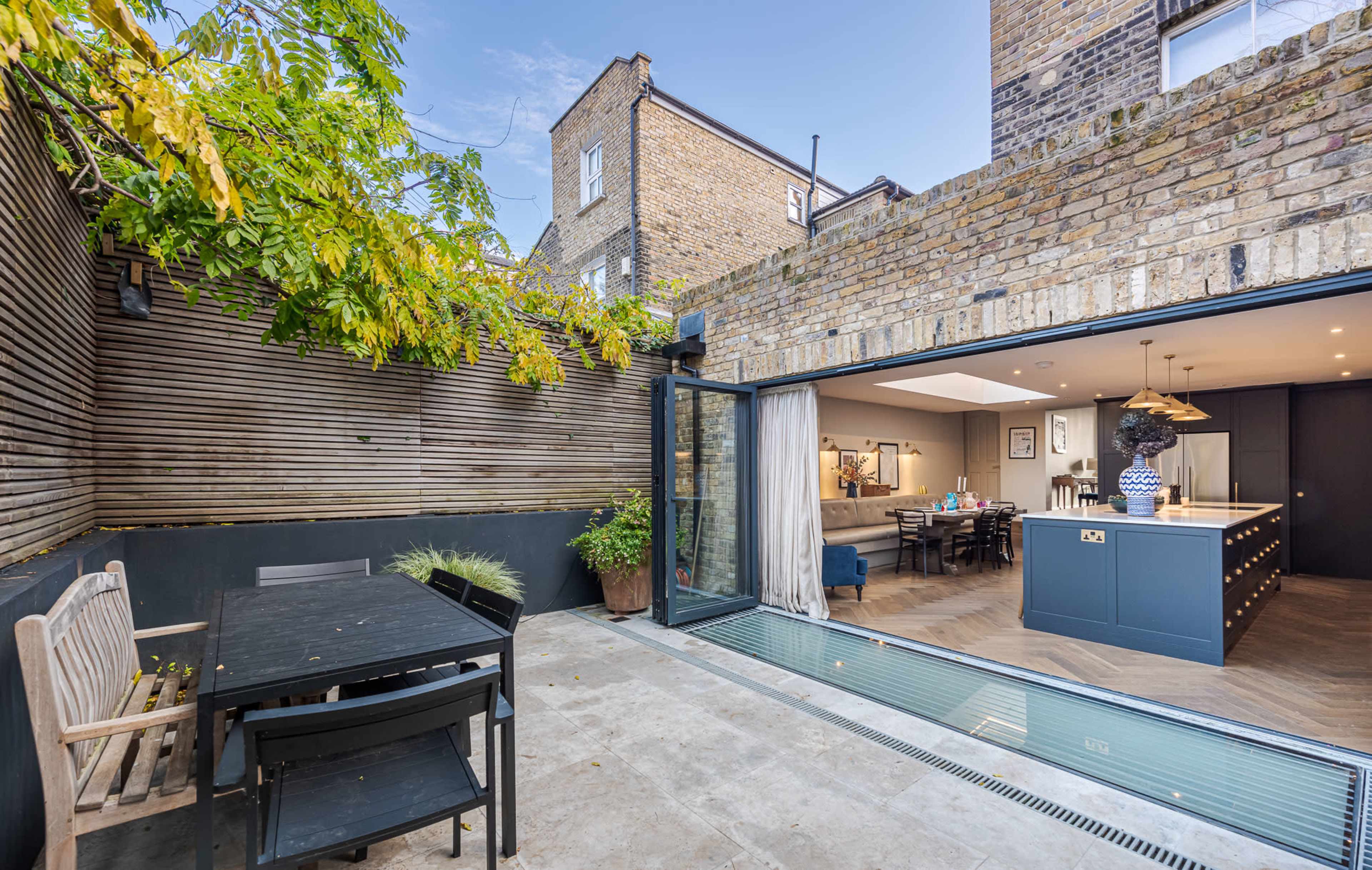 A modern outdoor dining area with a wooden table and chairs is connected to an open-plan kitchen and living space through large glass doors.