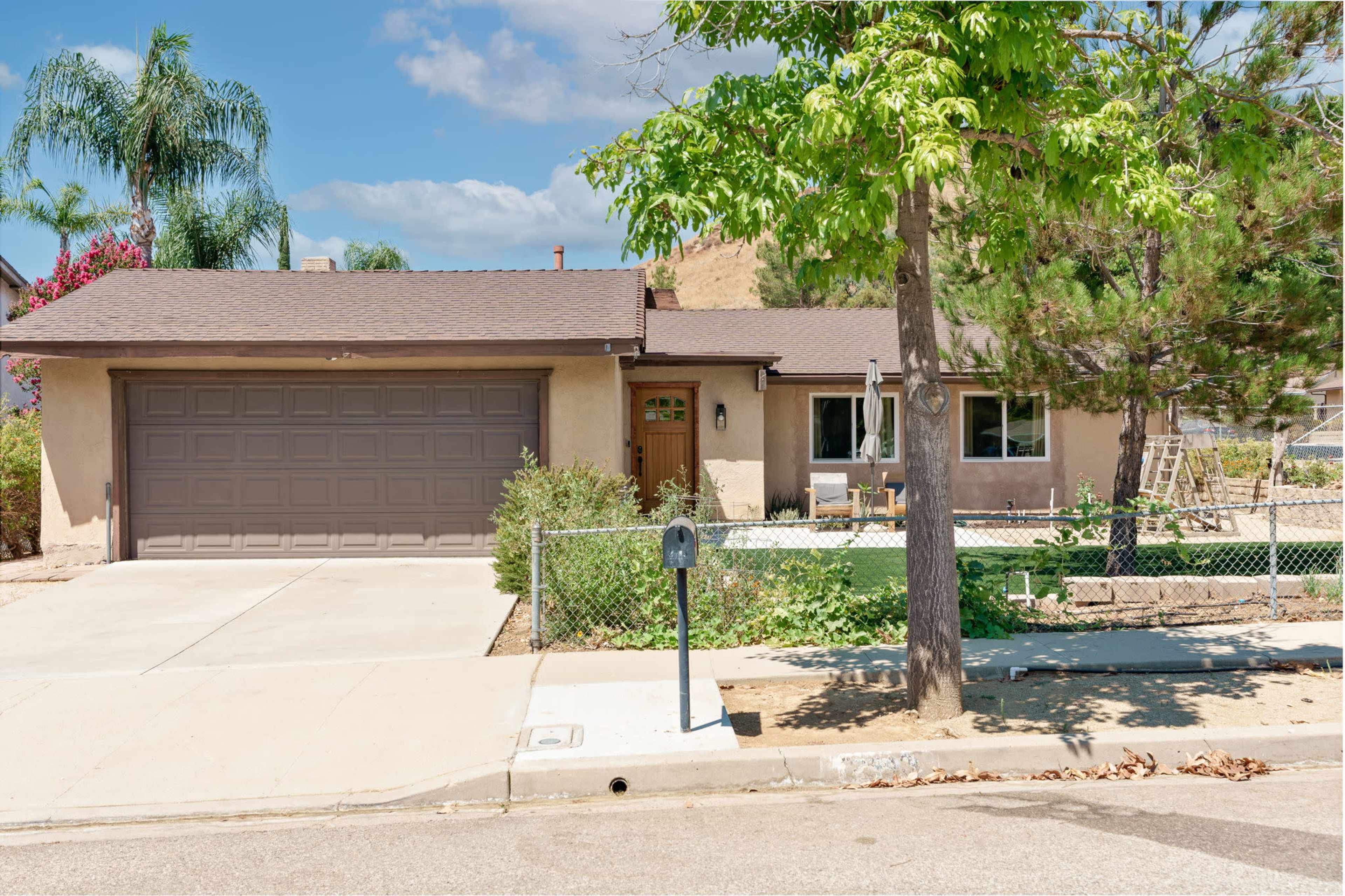 The image shows a single-story house with a brown garage door, surrounded by shrubs and trees, located on a quiet street.