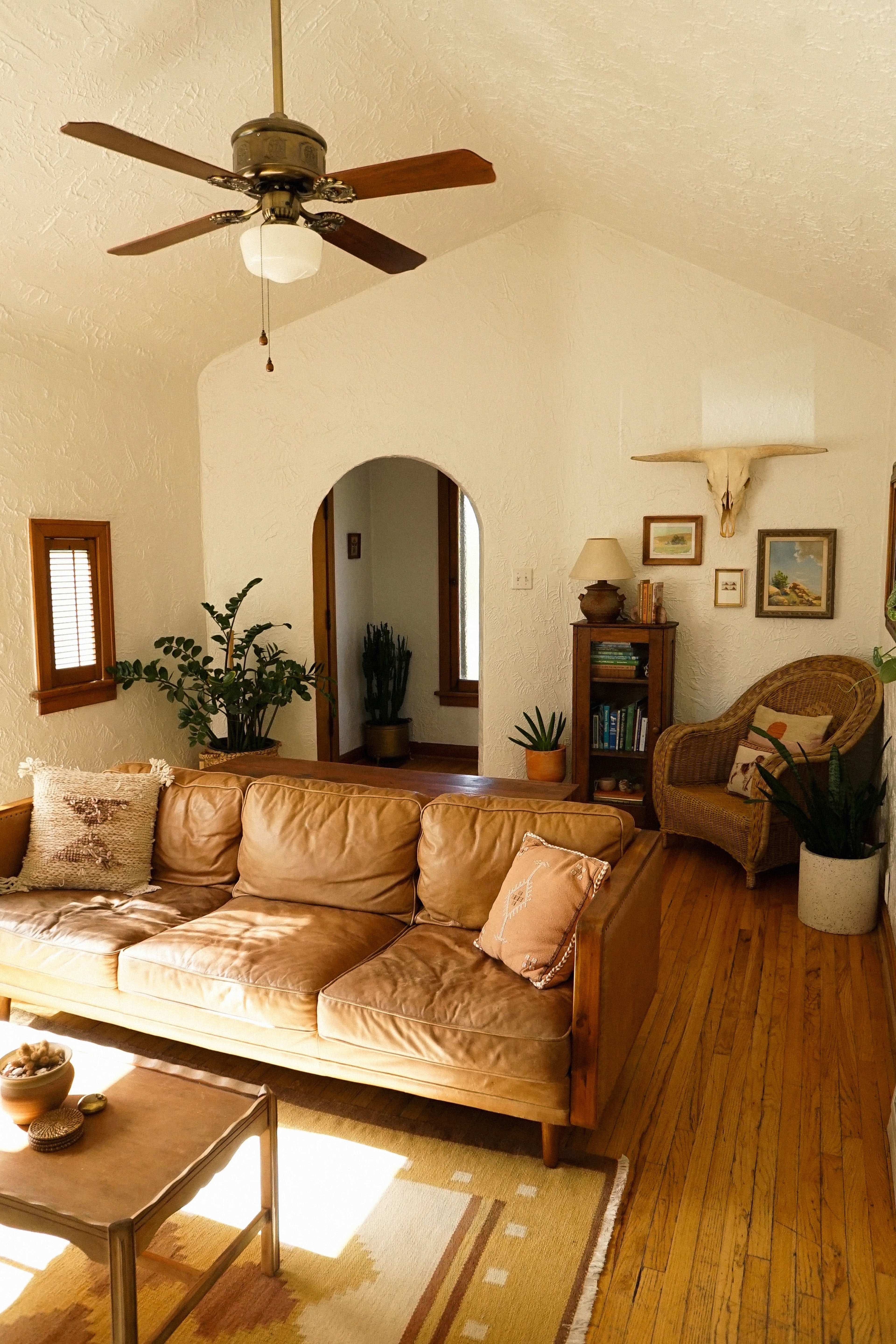 The image shows a cozy living room featuring a leather sofa, a wooden coffee table, houseplants, and a ceiling fan, with an arched doorway leading to another room.