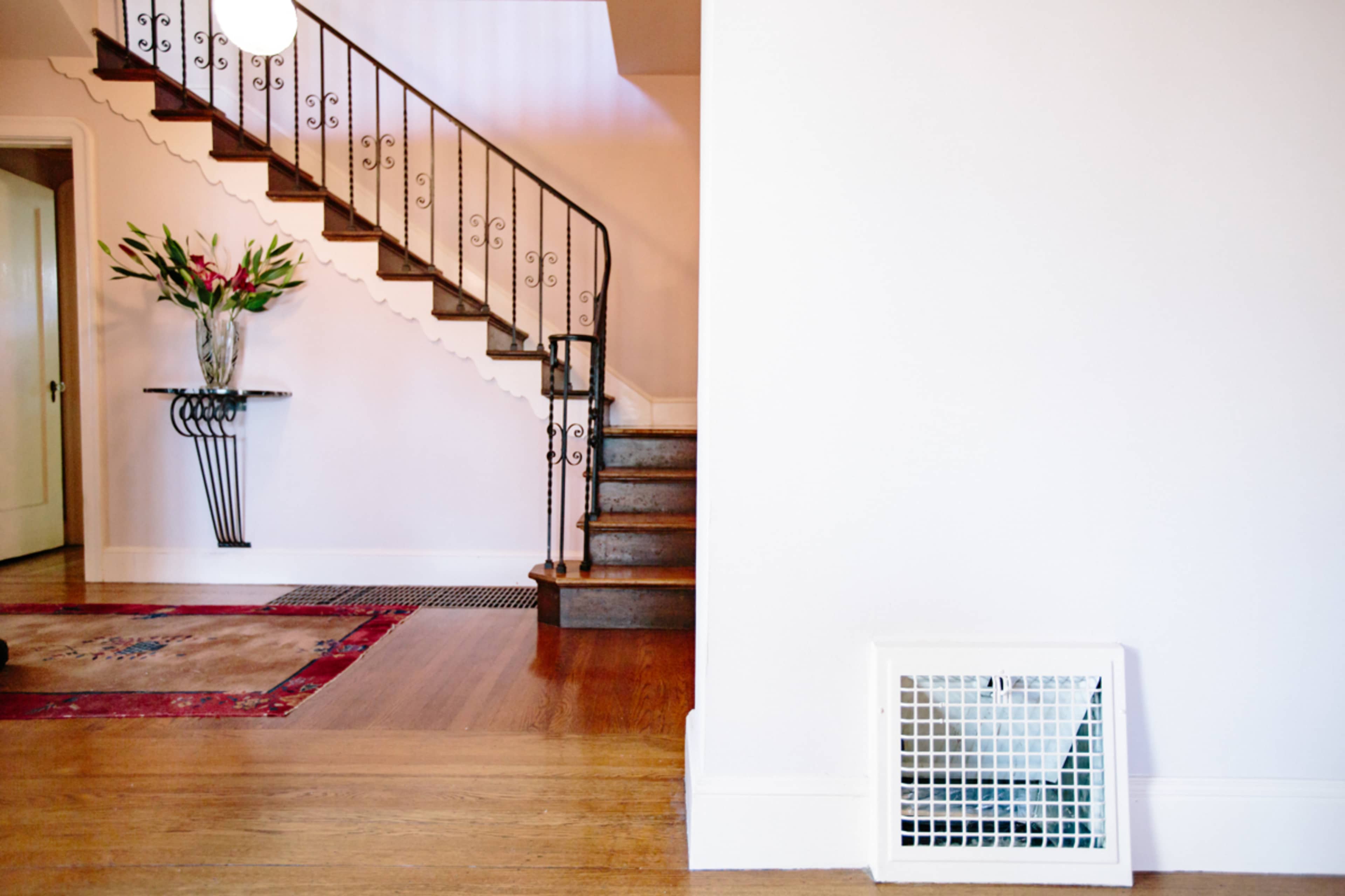 The image shows a clean entryway with a staircase on the left, a decorative table with a vase of flowers, and a vent on the right side.
