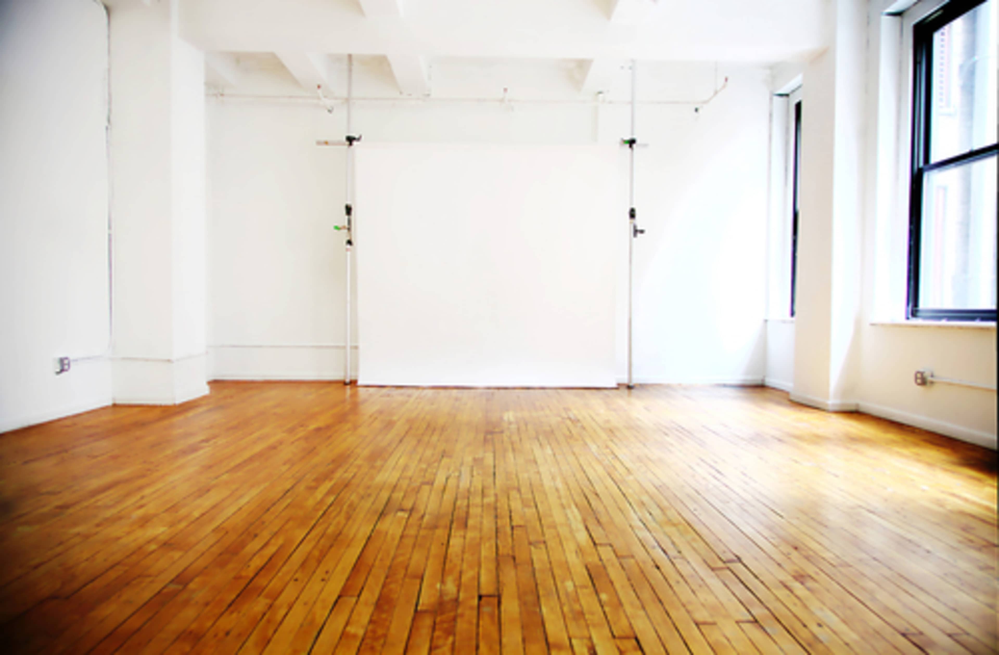 A bright, empty studio space features polished wooden floors and a blank white backdrop against the wall.