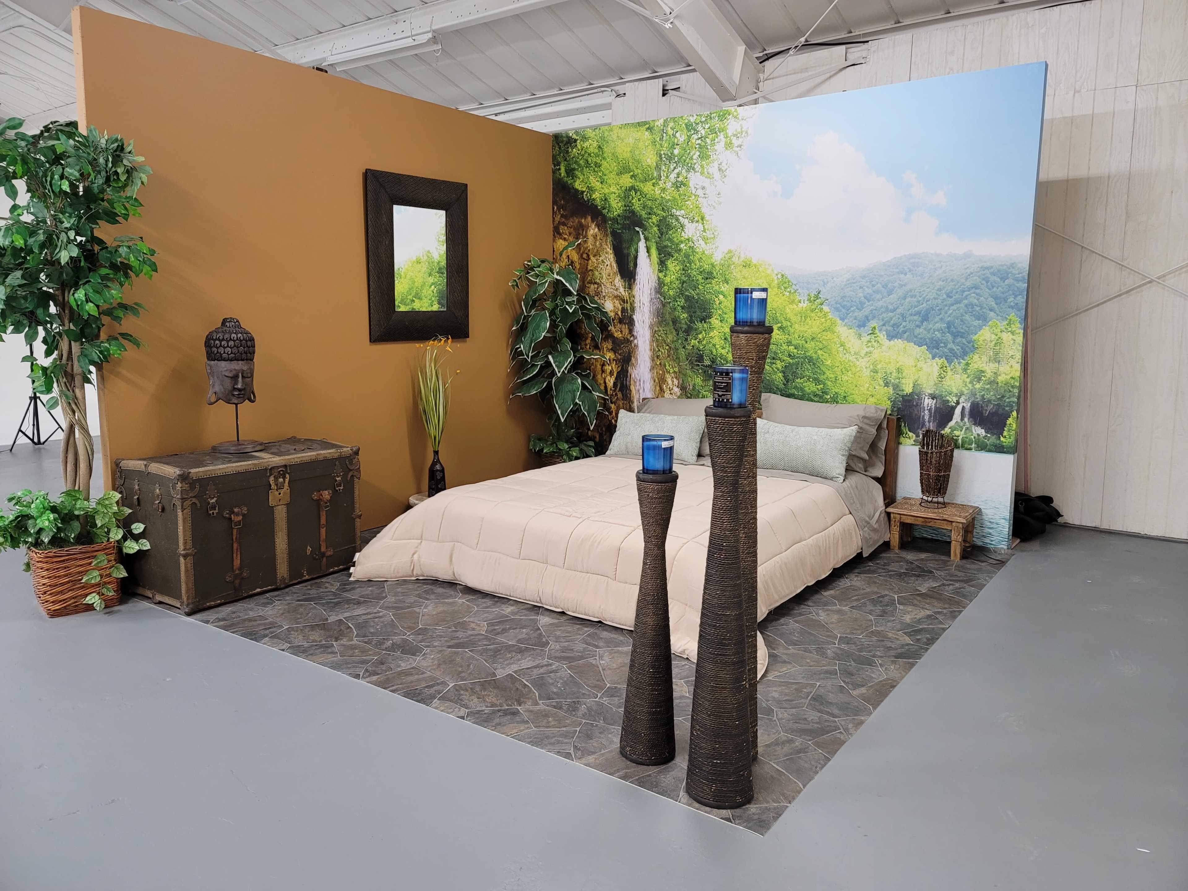 A staged bedroom scene featuring a bed with a beige quilt, two tall decorative stands with blue candles, a vintage trunk, and a backdrop of a natural landscape with a waterfall.